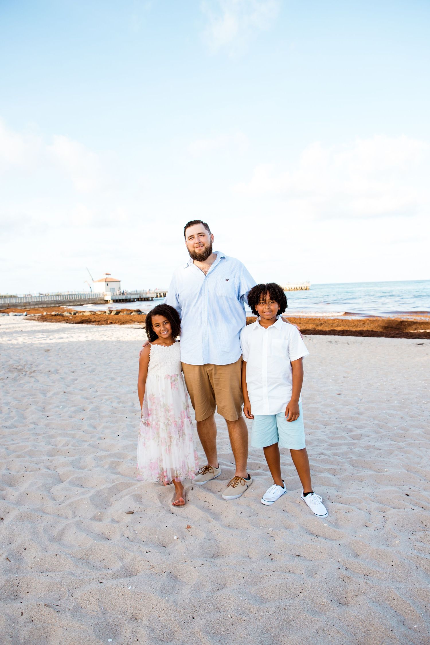 Three people pose for a portrait on a sandy beach with ocean waves and clear blue sky in the background.