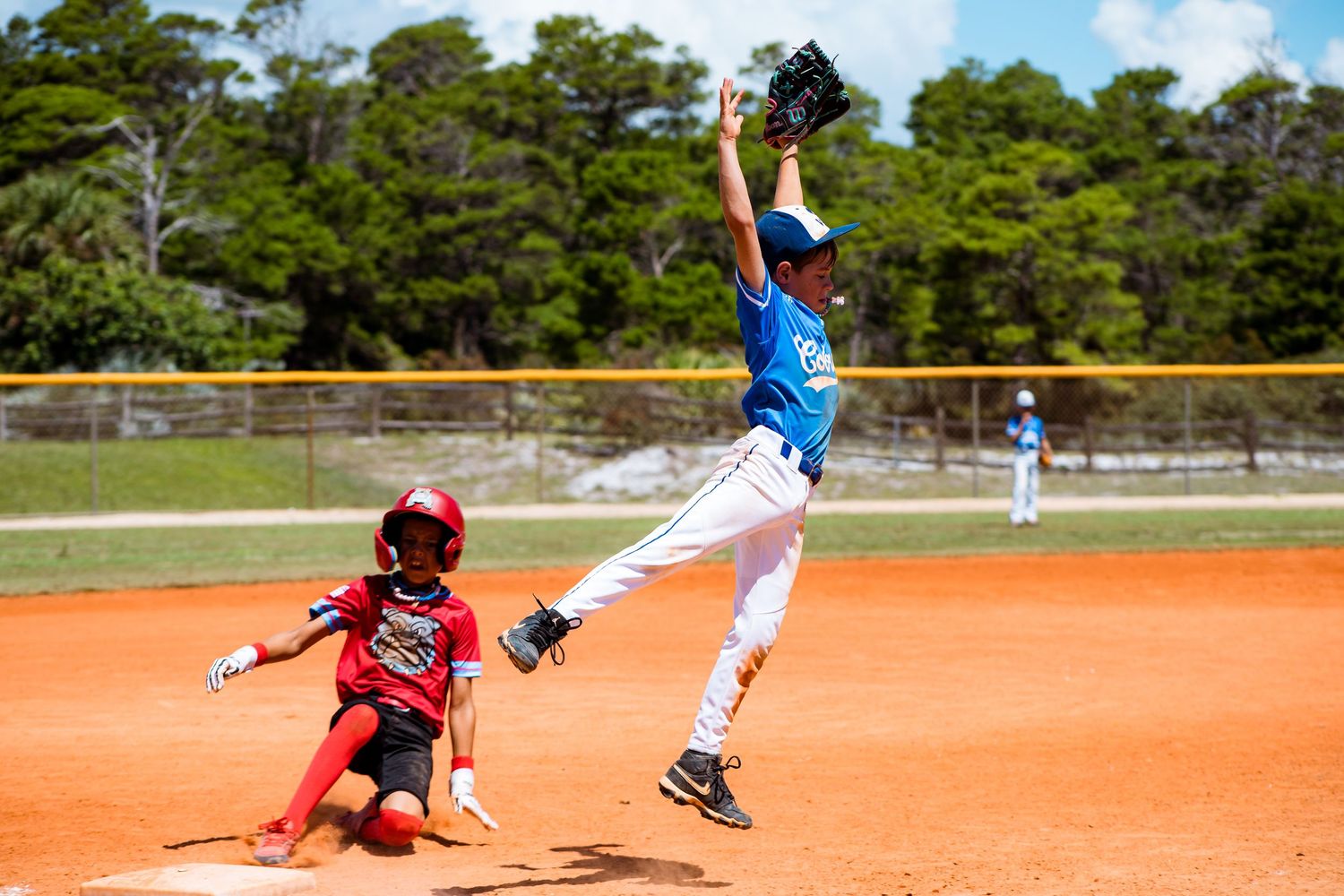 Baseball player in blue jersey leaps high to catch ball as runner slides into base.
