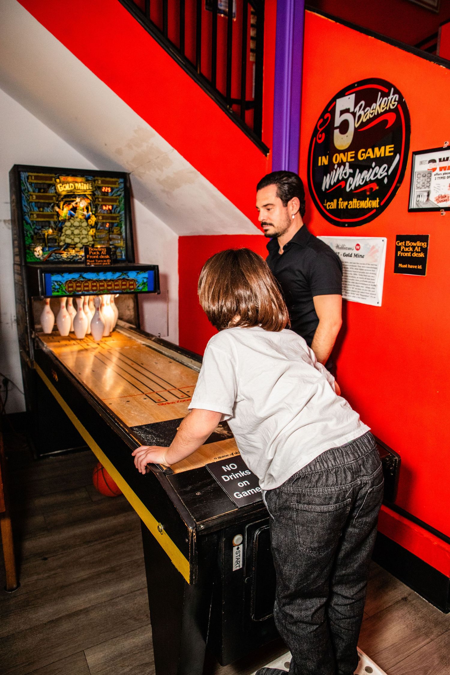 Child plays skee-ball arcade game with adult supervisor at a vintage indoor amusement venue.