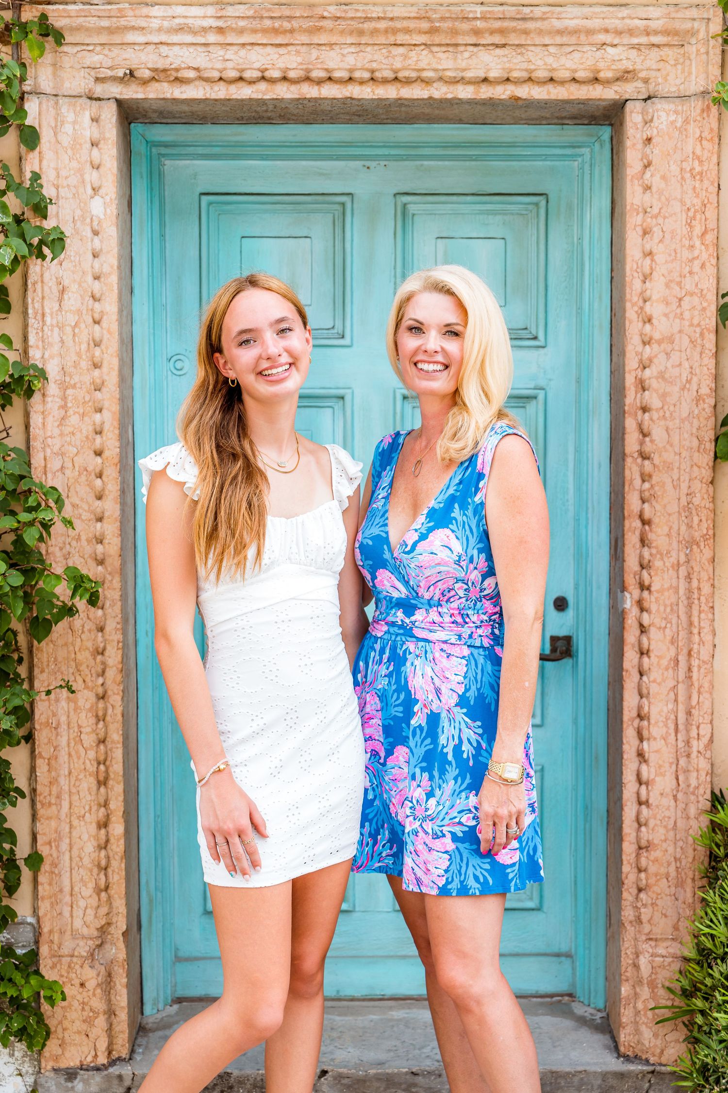 Two women pose in summer dresses in front of a turquoise double door with stone frame.