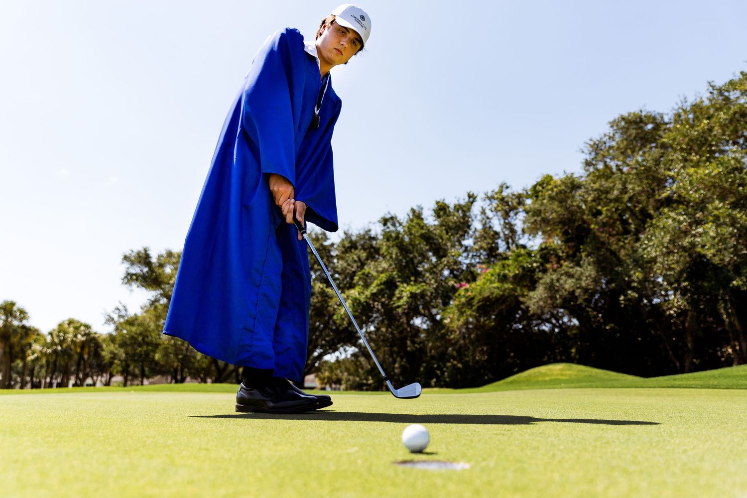 A golfer wearing a bright blue graduation gown prepares to putt on a sunny golf course.