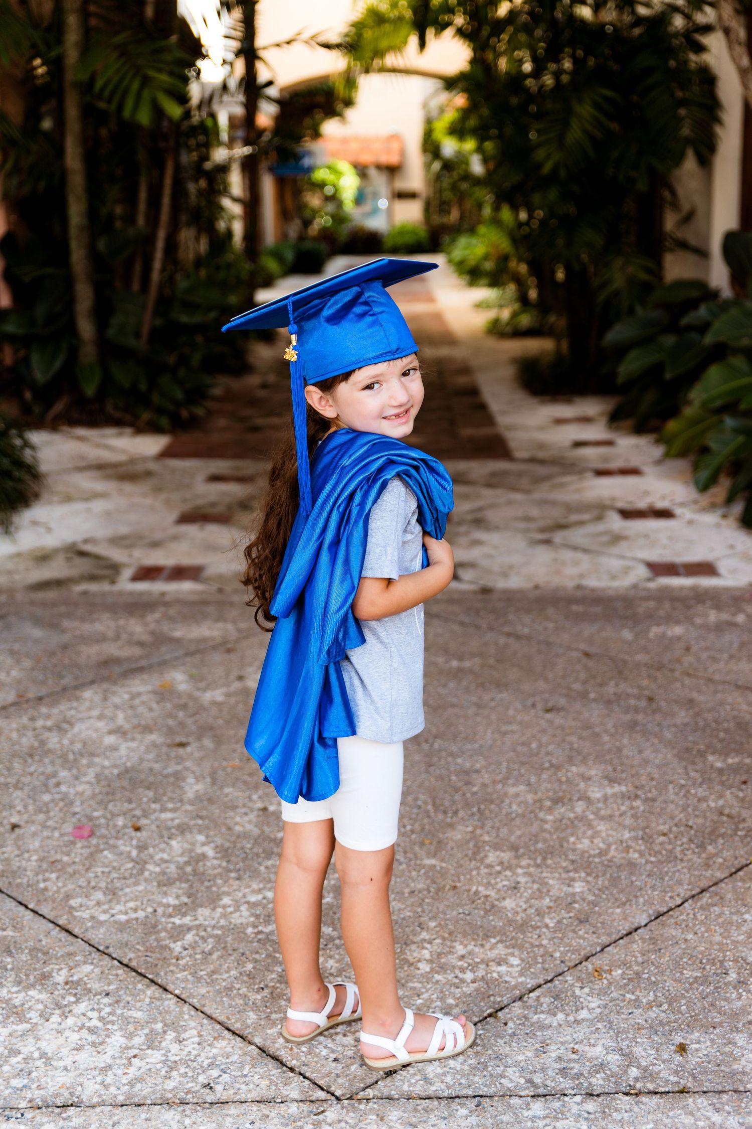 A child in white shorts and gray shirt smiles while wearing a blue graduation cap and gown on a stone walkway.