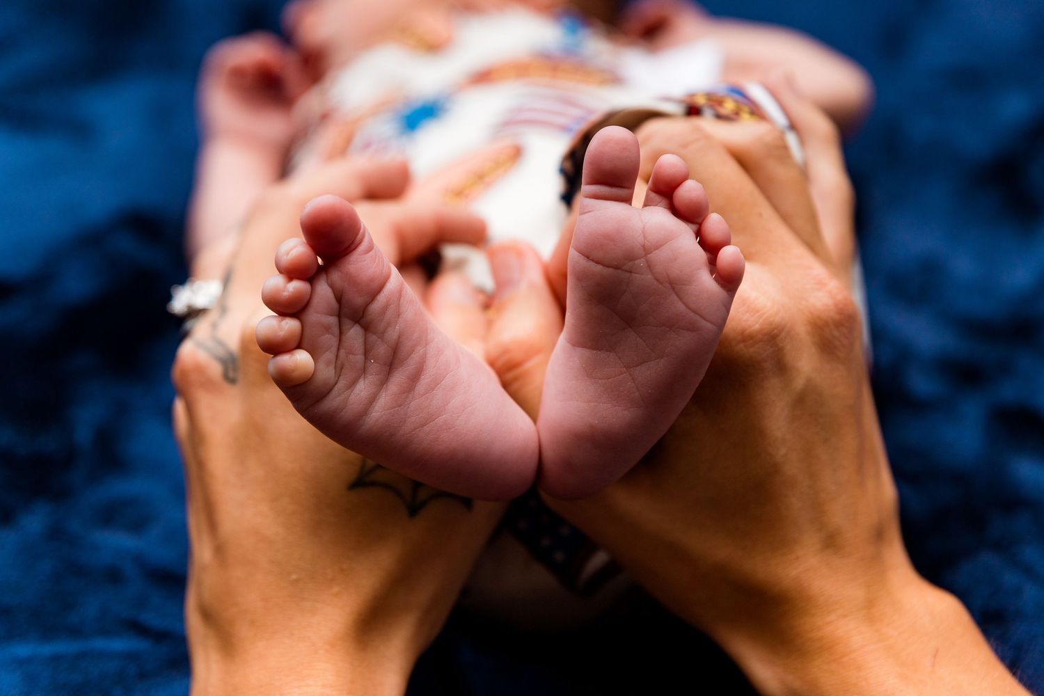 Adult hands gently cradle tiny newborn baby feet against a dark blue background.