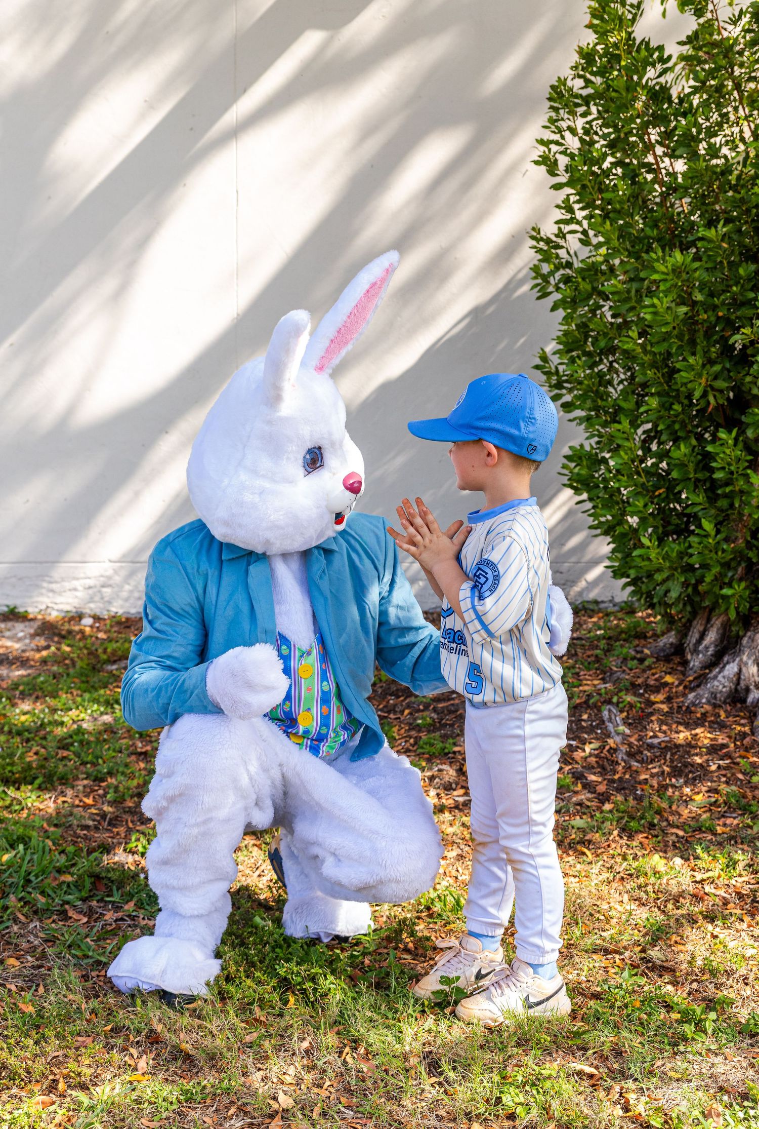 Child in baseball uniform giving high five to Easter Bunny mascot crouching on grass outdoors.
