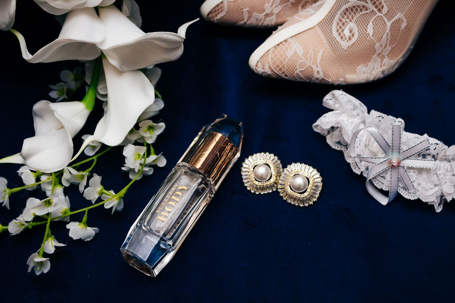 Bridal accessories including flowers, earrings, and perfume arranged on dark blue fabric.