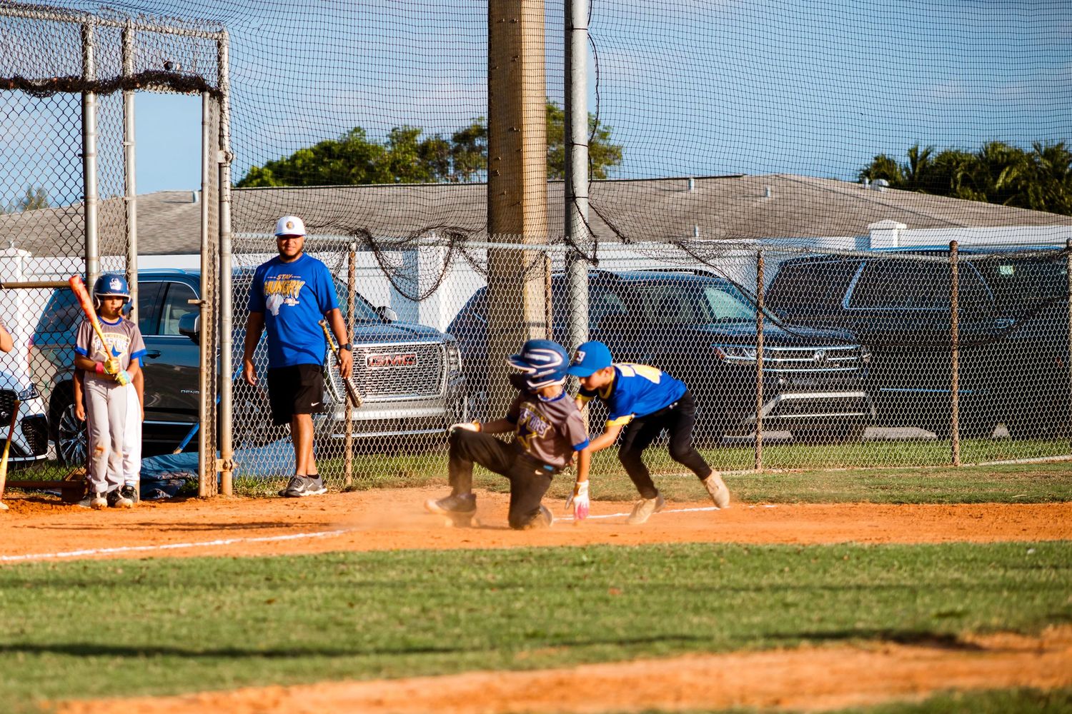 Baseball players in blue uniforms compete during a game on a dirt infield with spectators watching from the sidelines.