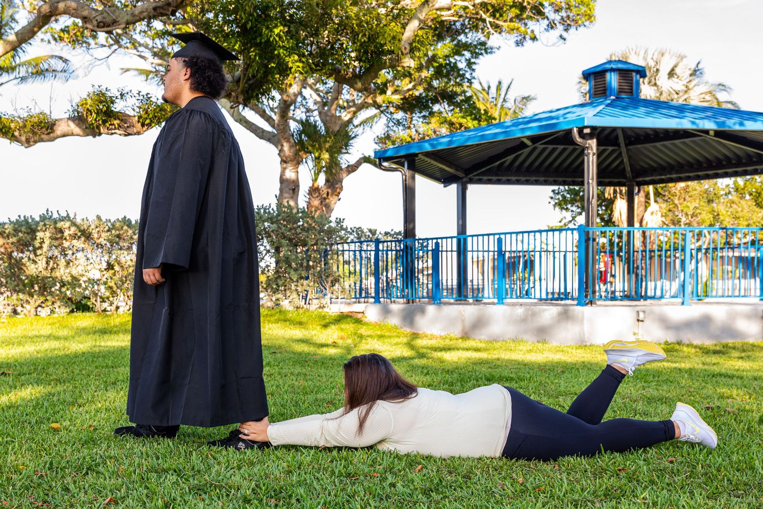 Graduate in cap and gown stands on grass while photographer lies down for low-angle shot near blue gazebo.