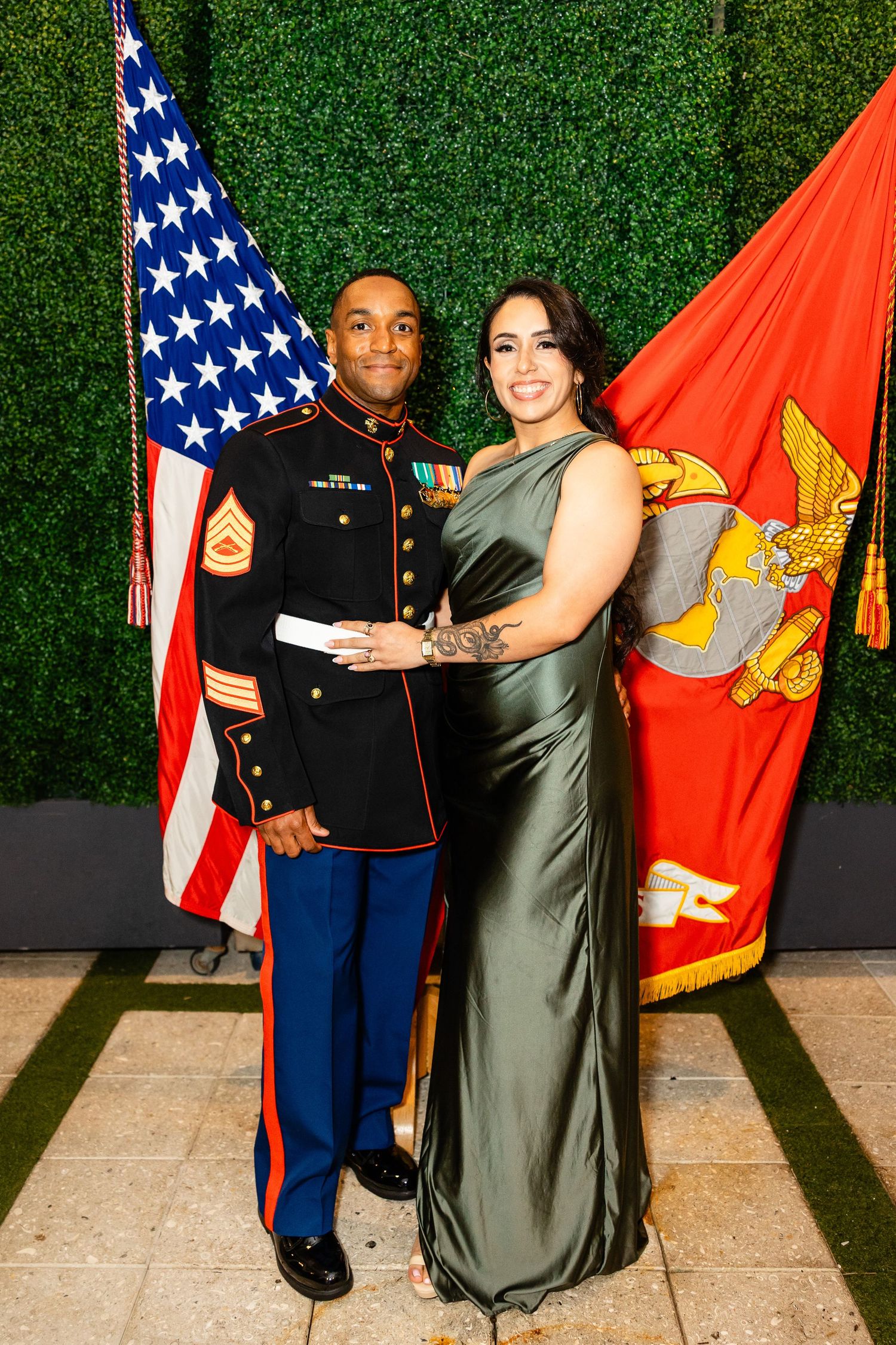 Marine in dress blues uniform stands with companion in elegant sage green gown against hedge wall.