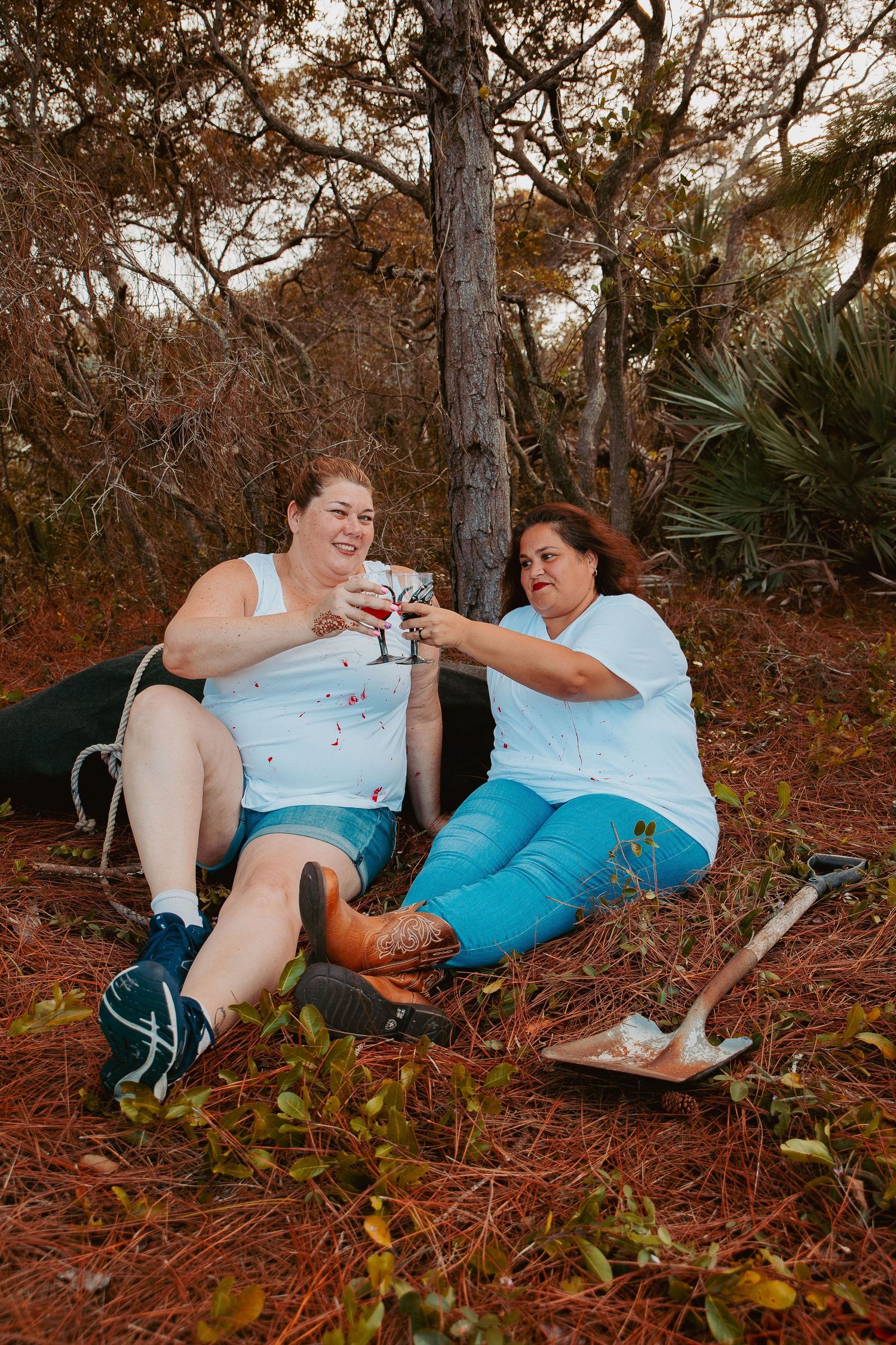 Two people in casual summer clothing share a toast while sitting on the ground in a wooded outdoor setting.