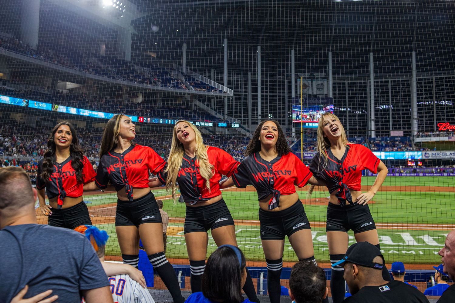 Baseball cheerleaders in blue uniforms perform a dance routine along the sidelines of a stadium field.