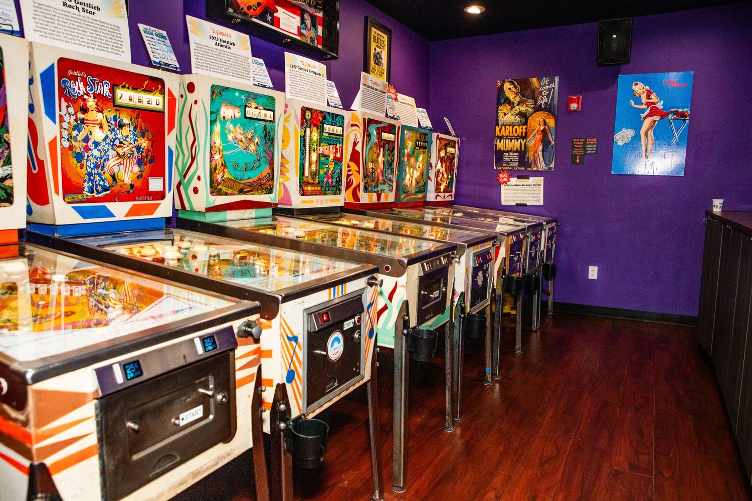 A row of vintage pinball machines lines the walls of a colorful arcade game room with purple walls and wood floors.