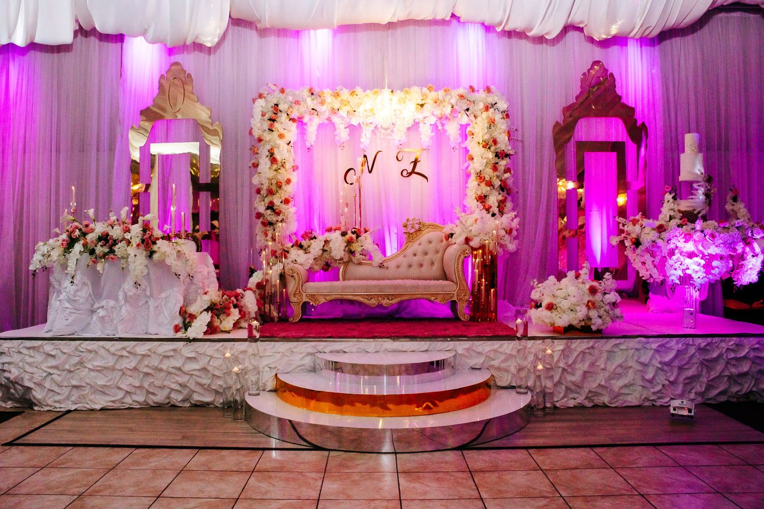 Grand wedding stage setup with floral arrangements, loveseat, and pink ambient lighting.