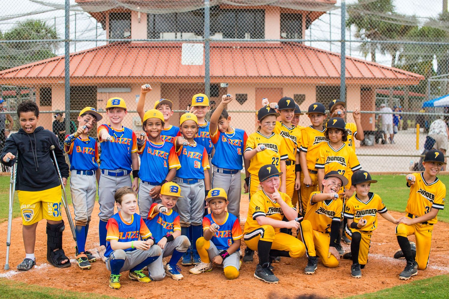 Two youth baseball teams in blue and yellow uniforms pose together for group photo on field.