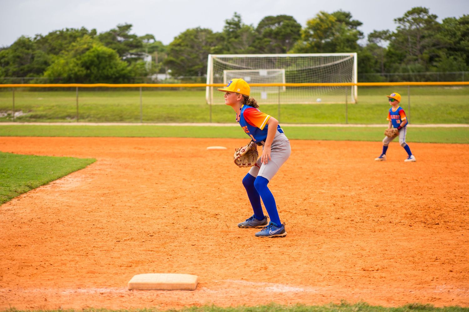 A baseball player in bright blue and orange uniform stands ready at their position on the infield.