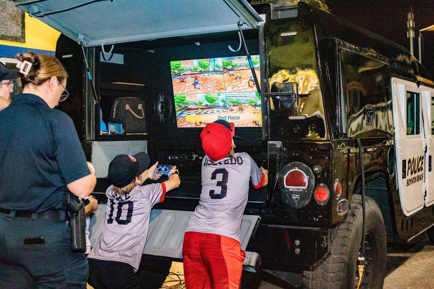 Young players in baseball jerseys play a video game at an outdoor gaming station.