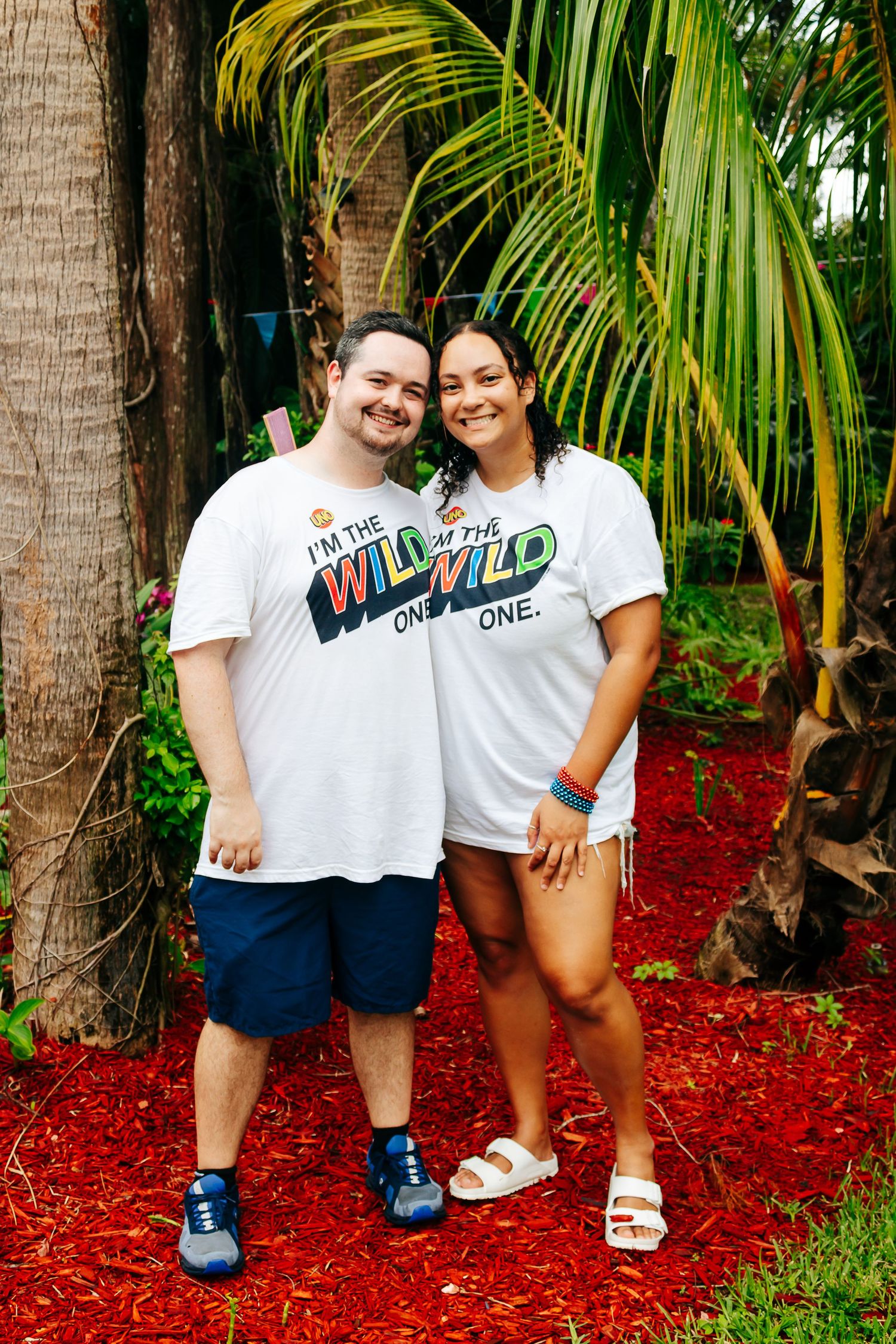 Couple wearing matching white graphic t-shirts posing in front of palm trees.
