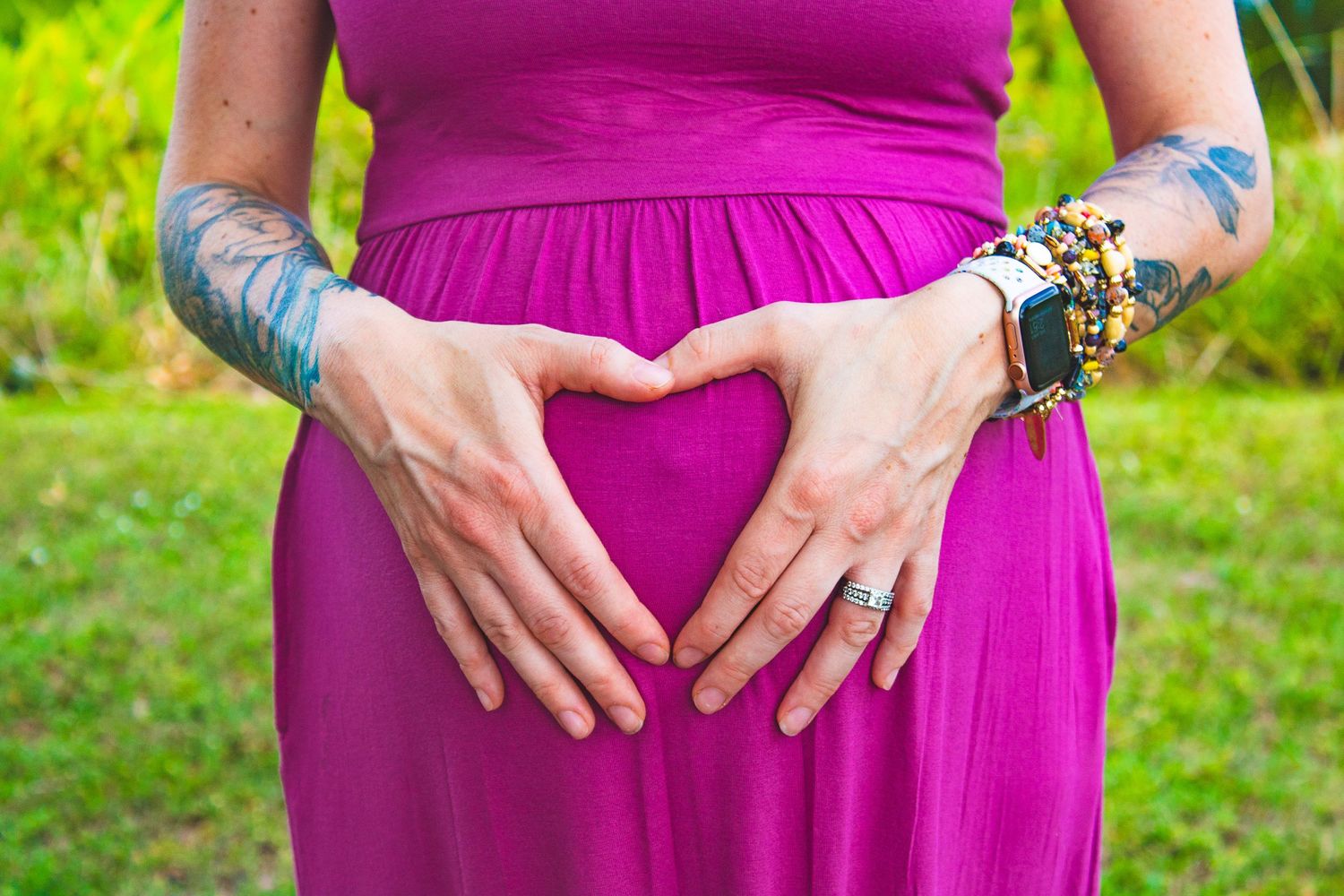 Hands forming heart shape over magenta maternity dress with visible tattoos and bracelets.