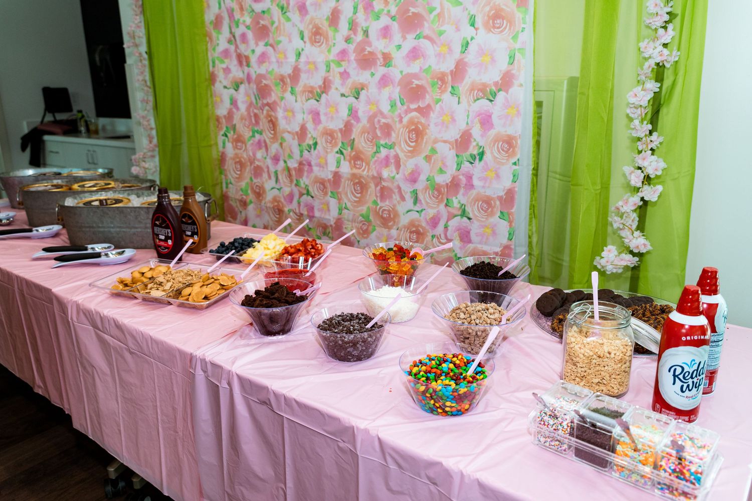 Candy and dessert buffet table with pink tablecloth features colorful toppings against a floral backdrop.