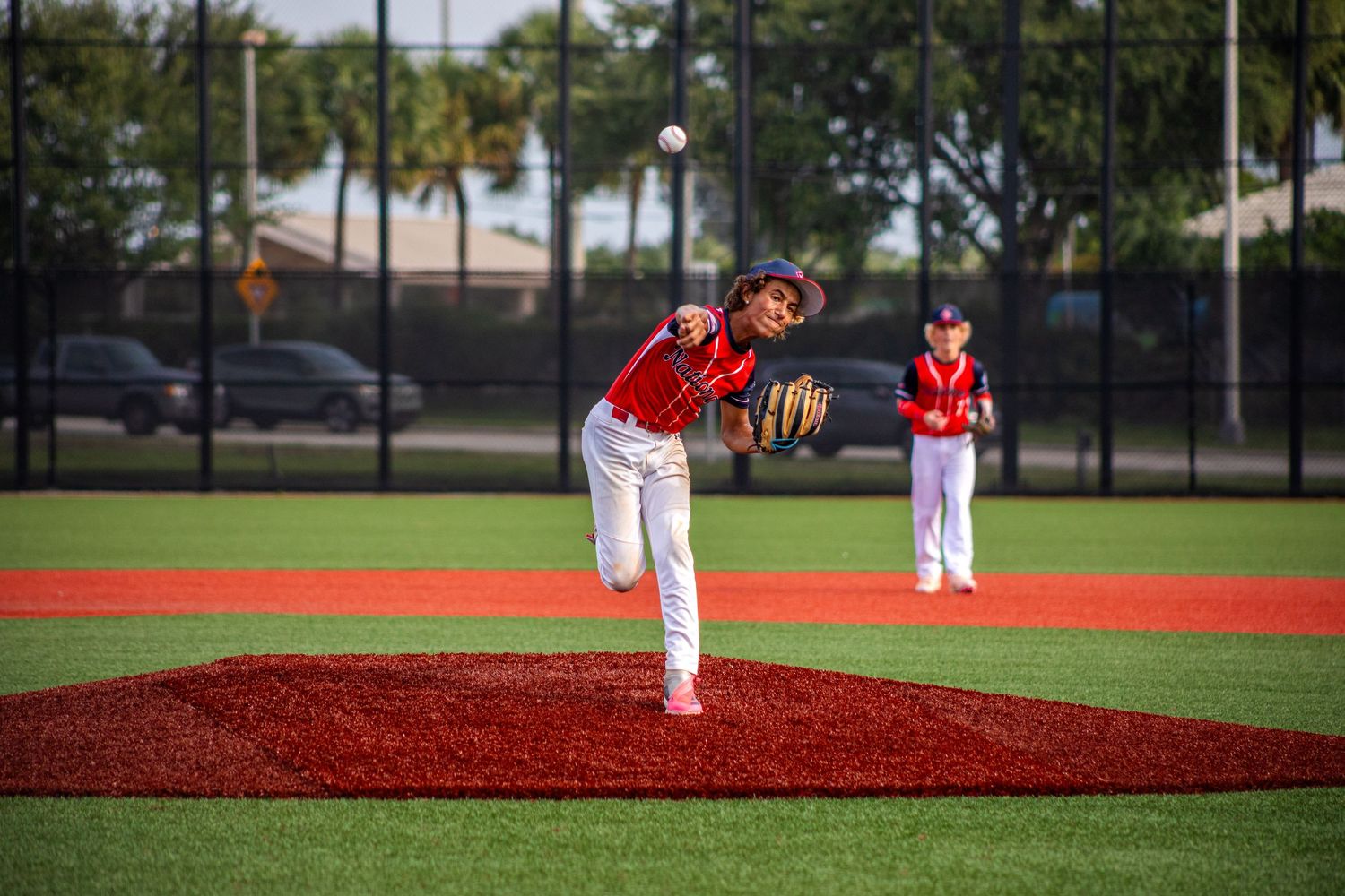 Young baseball pitcher in red uniform throws ball on mound during youth game outdoors.