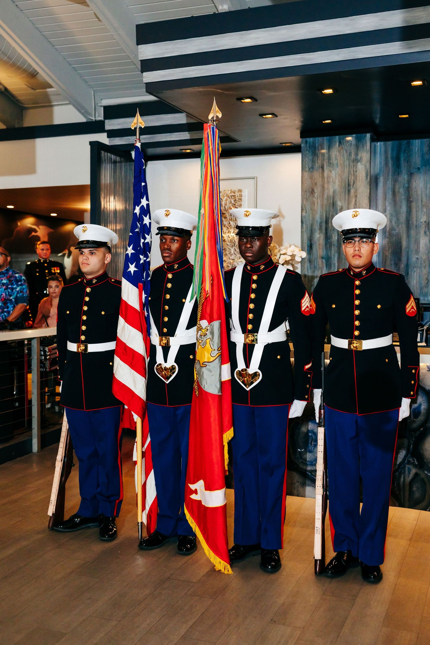 Marines in dress blues uniforms stand at attention in formation with American and Marine Corps flags.