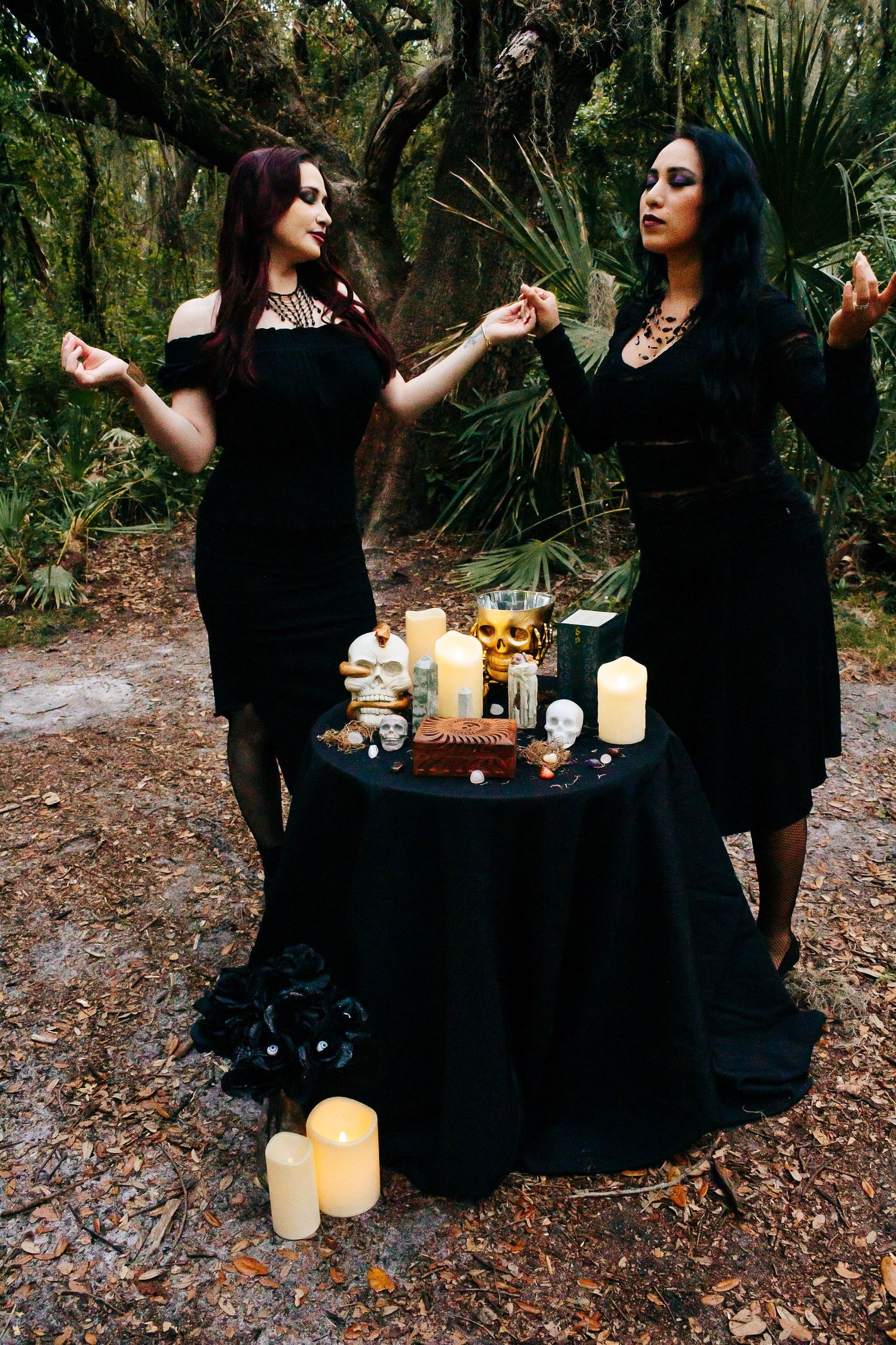 Two figures in black dresses perform a ritual around a table with candles and crystals in a mystical forest setting.