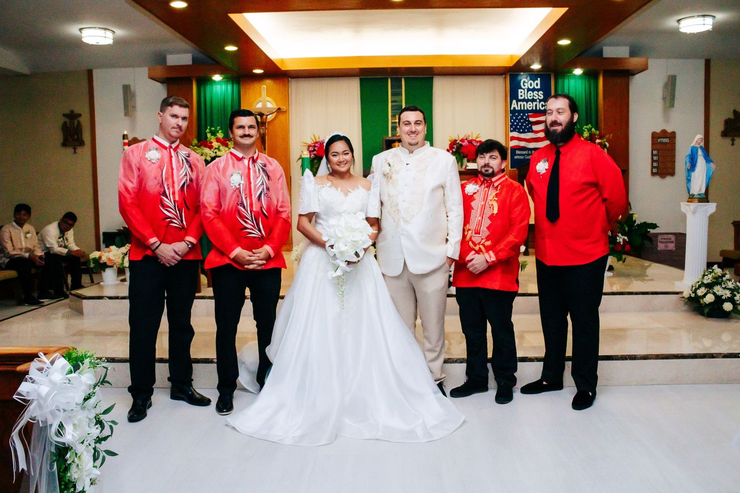 Wedding party members in red attire pose with bride and groom at church altar.