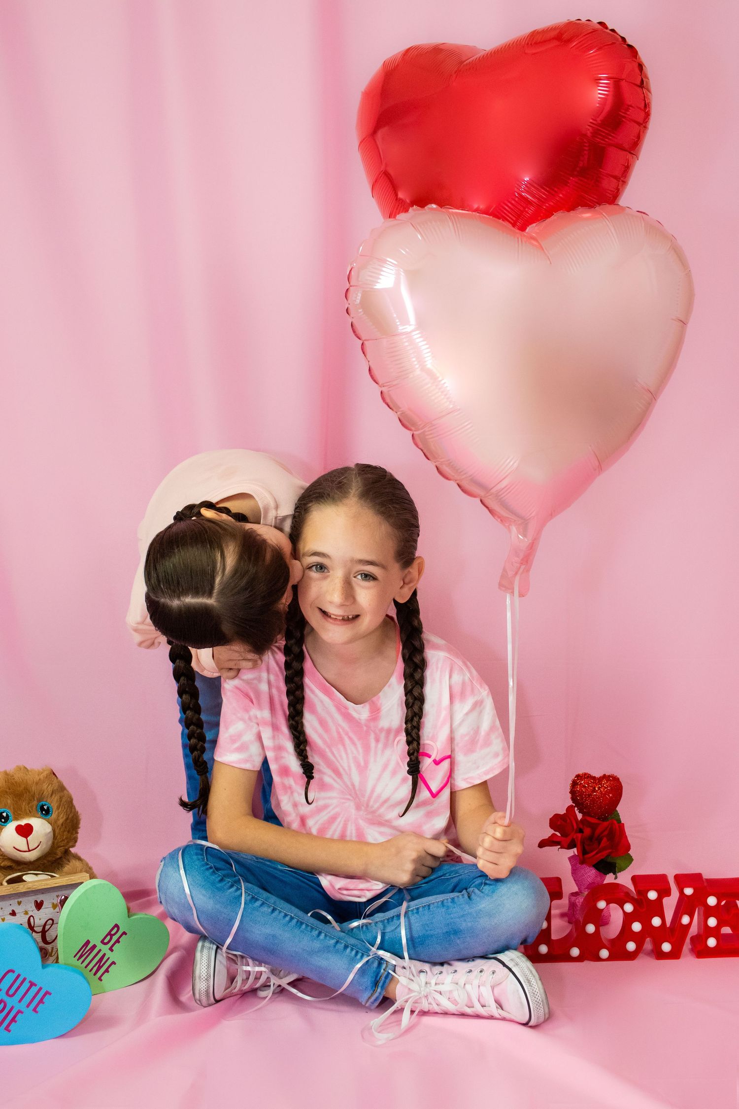 Two children embrace while sitting with heart balloons and presents on pink backdrop.