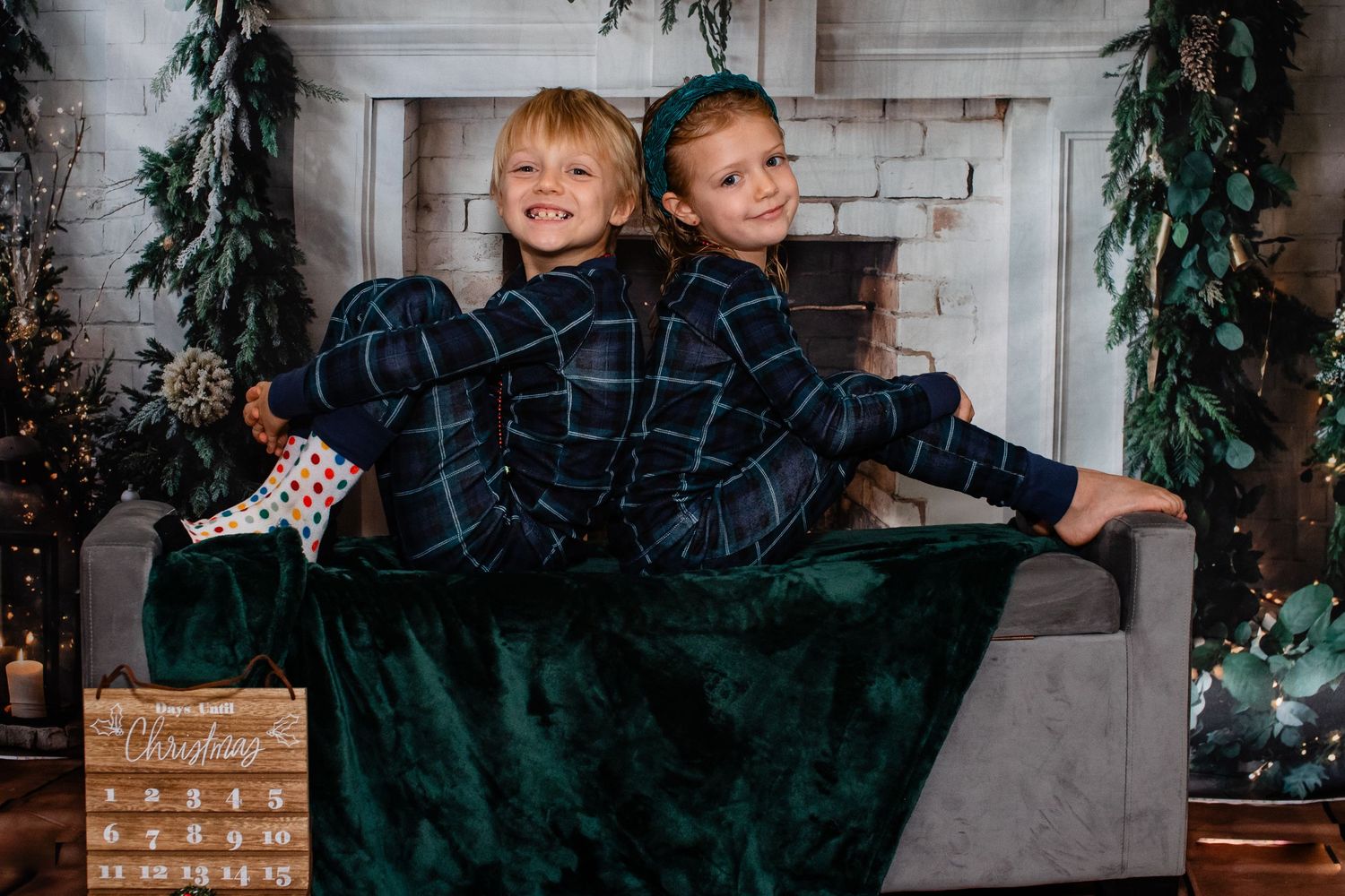 Two children in matching plaid pajamas sharing a cozy moment by a decorated fireplace during the holidays.