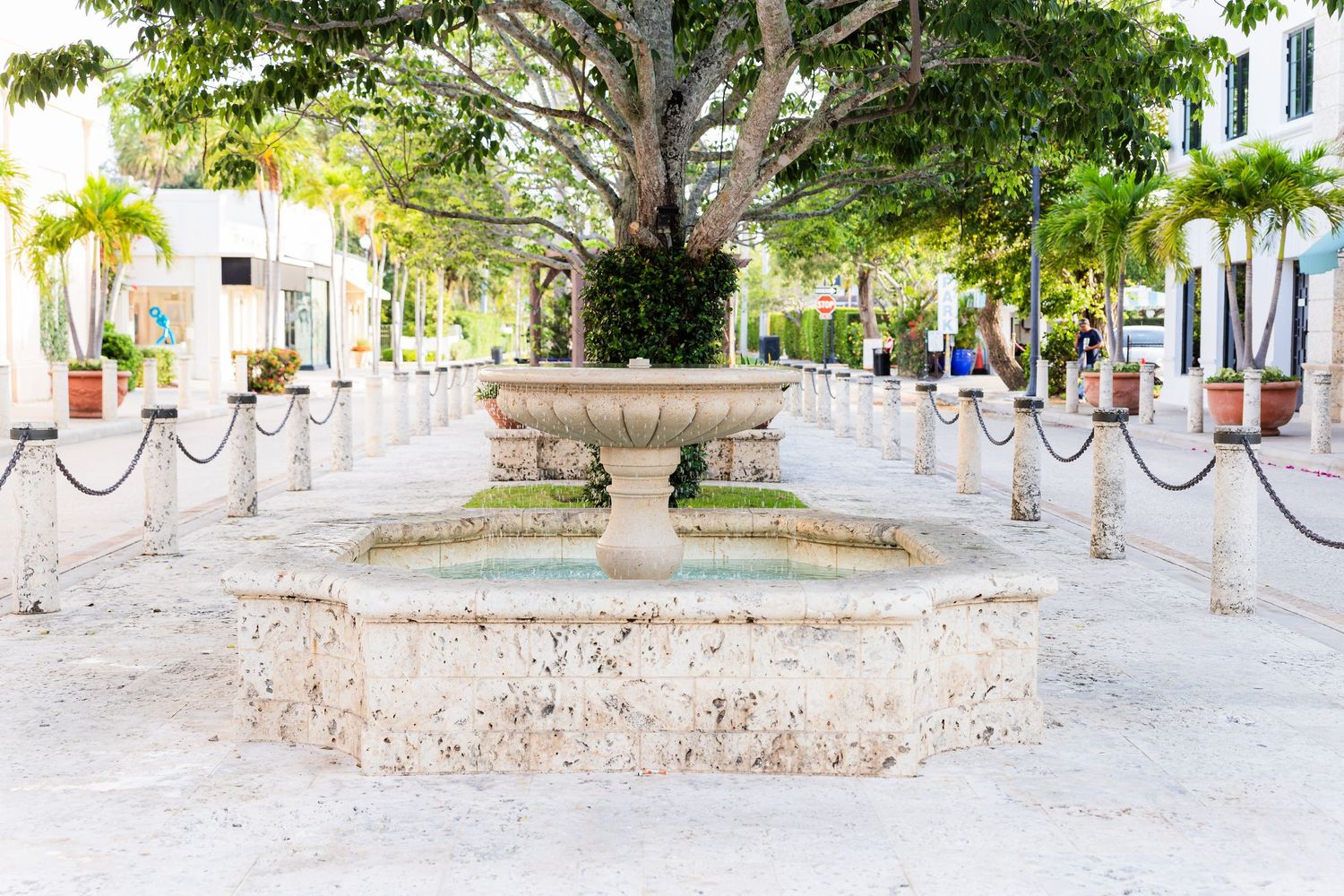 A stone water fountain stands in the center of a rope-lined walkway with green trees and white buildings in the background.