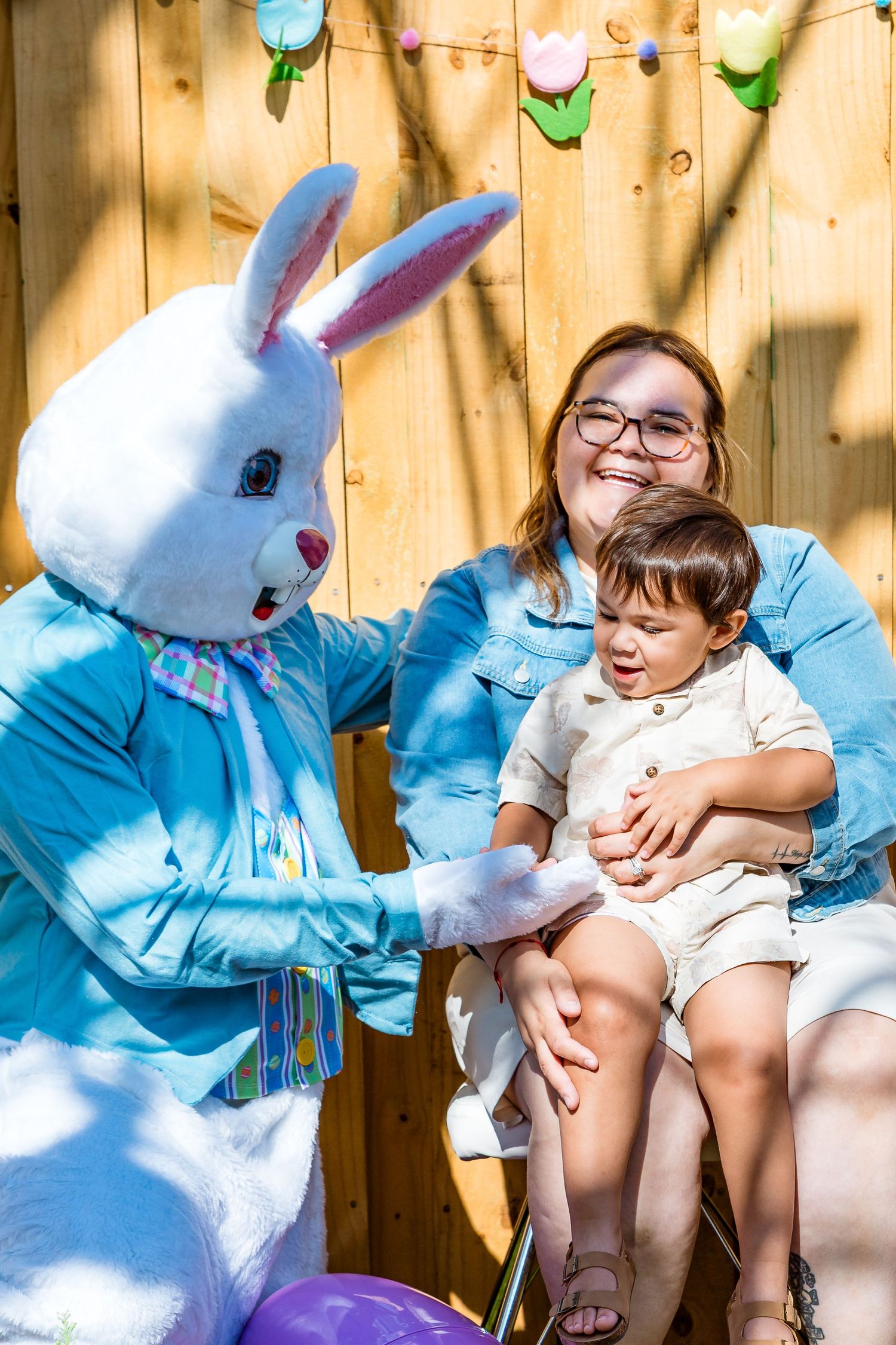 A person and child sit next to a large Easter Bunny costume character in front of a wooden fence with decorations.