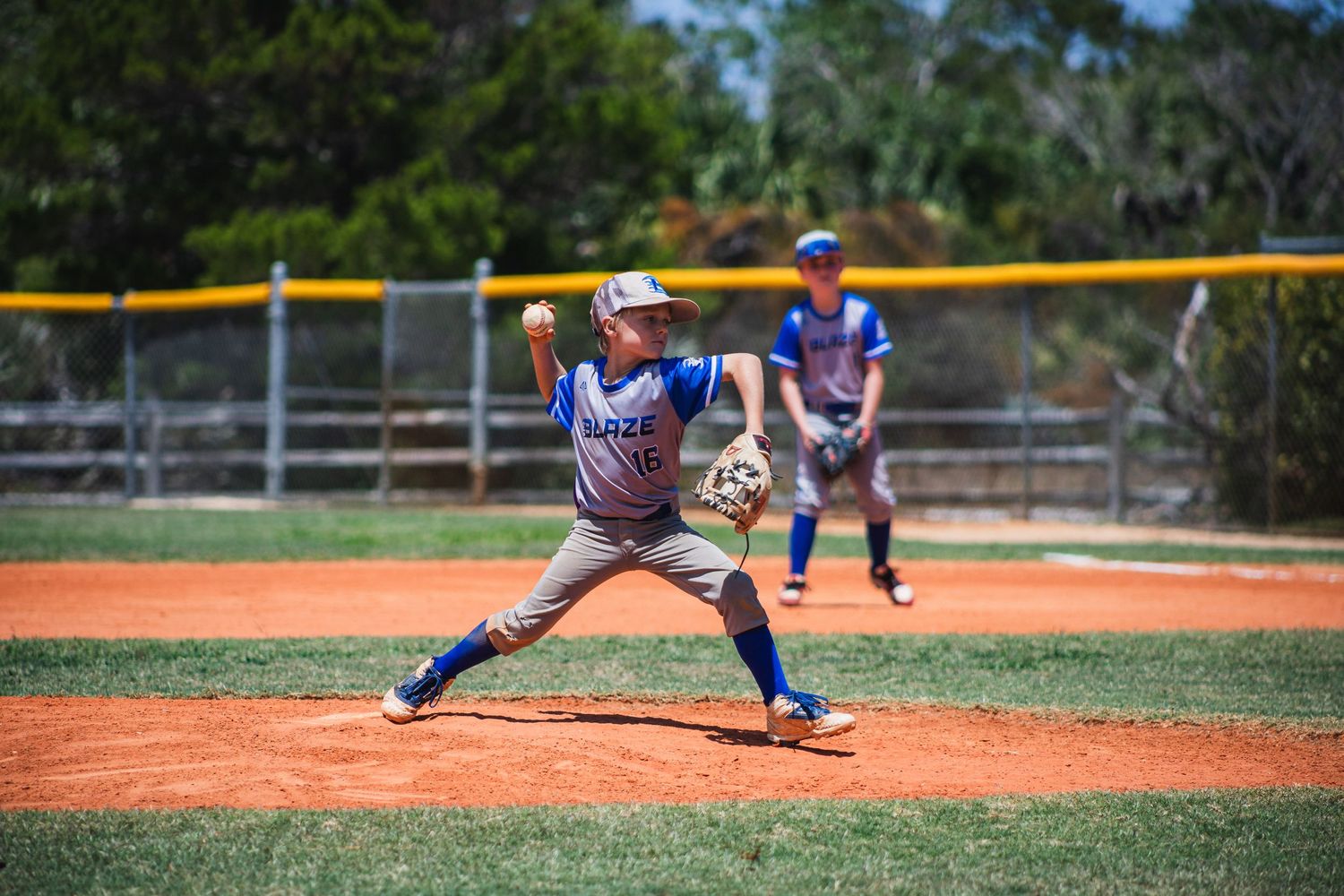 Baseball player in blue uniform pitches from the mound during a sunny game on red clay diamond.