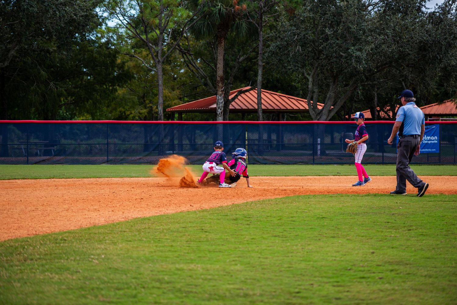 Baseball players in action during a game on a sunny day at a baseball field with red dirt infield and green grass.