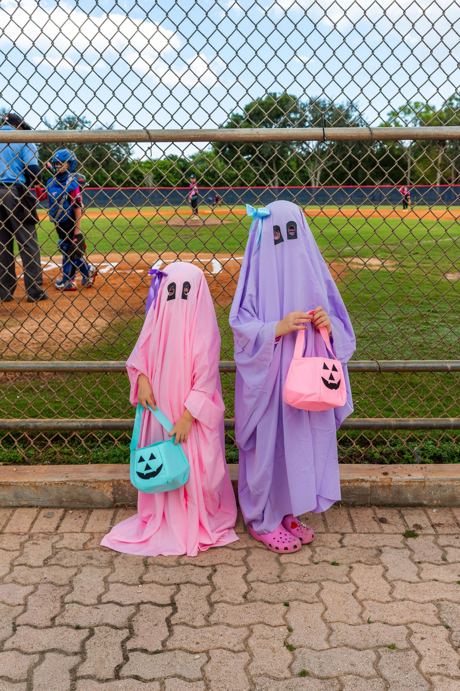 Two people dressed as colorful ghosts with Halloween bags watch a baseball game through a chain link fence at a ballpark.