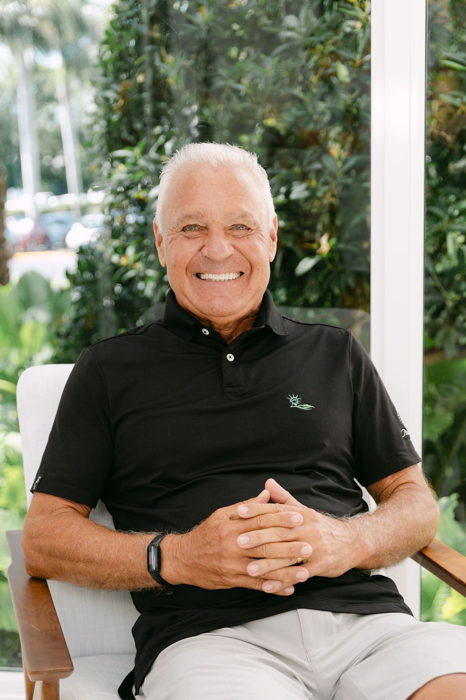 Smiling senior man with white hair sitting relaxed in a white chair, wearing a black polo shirt, outdoors with green foliage.