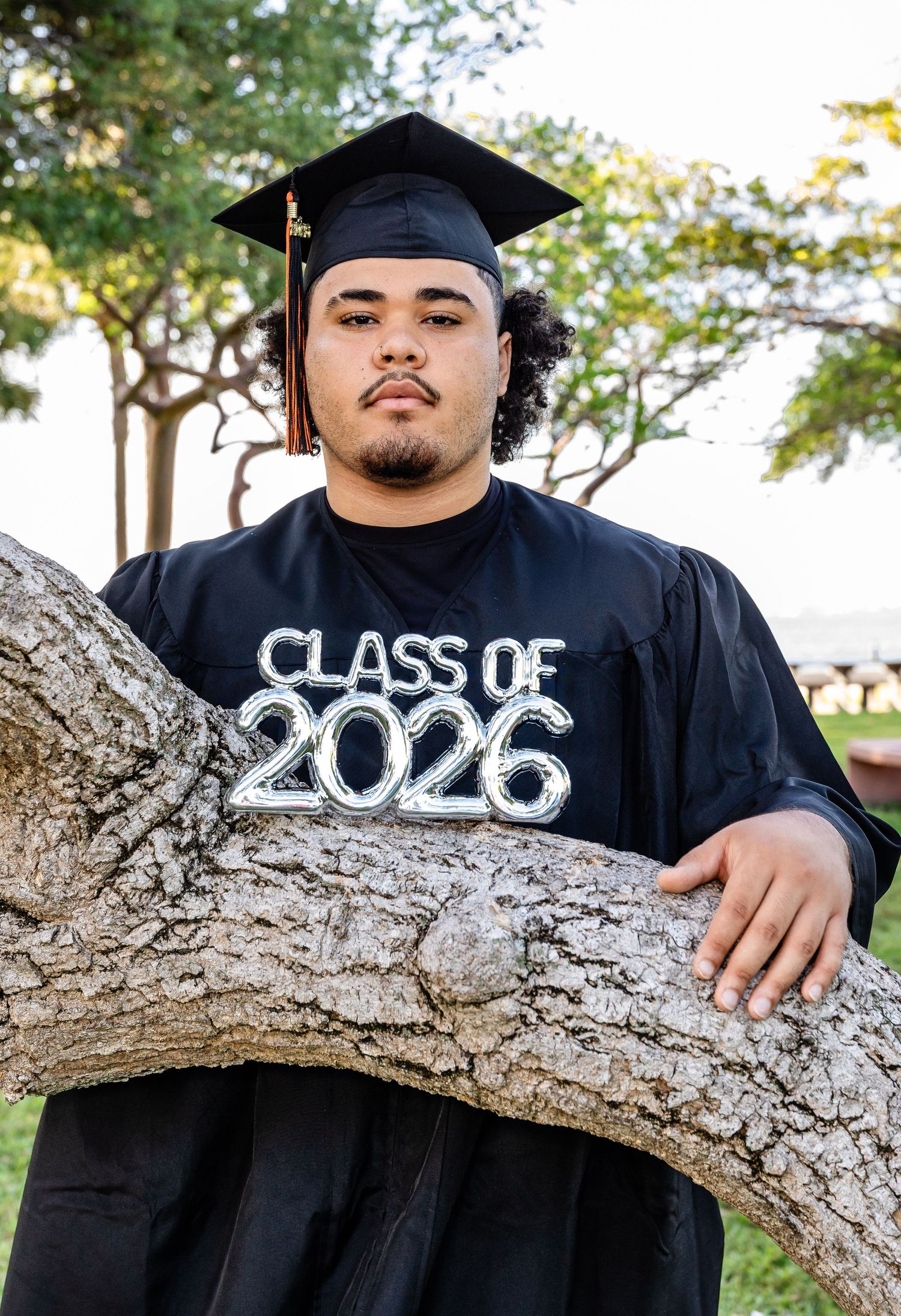 Young man in black graduation cap and gown with Class of 2026 emblem poses by a tree outdoors.