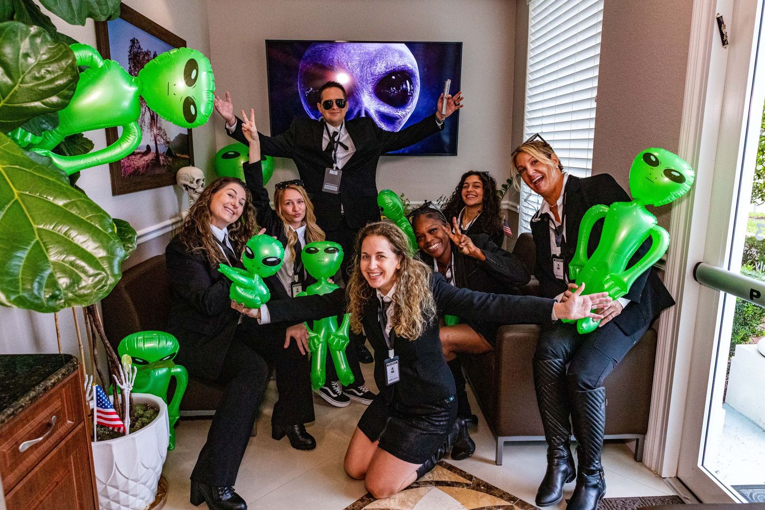 A group celebrates with green alien balloons and decorations at an indoor party gathering.