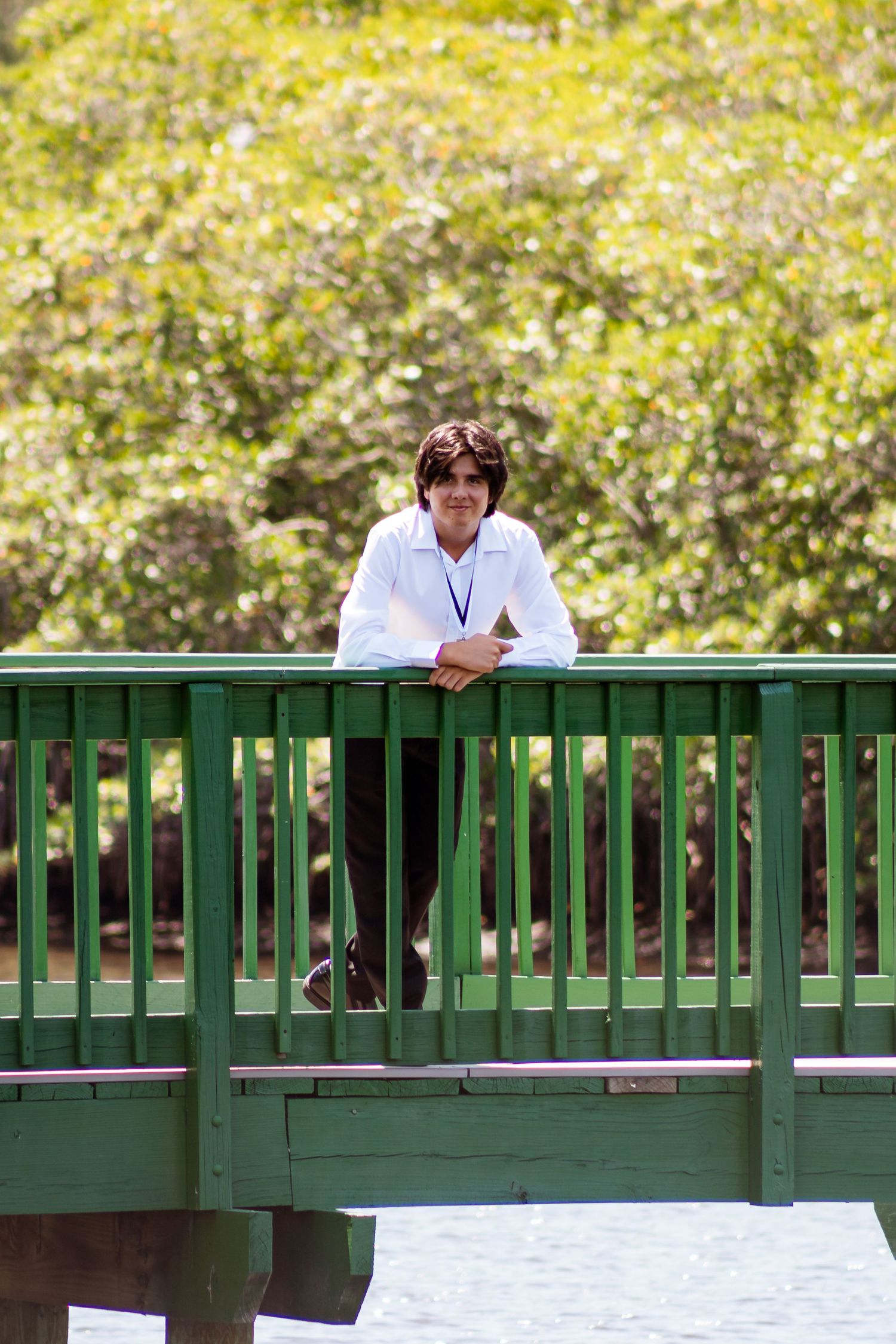Someone in business attire leans against a green railing on an outdoor bridge with sunlit foliage in the background.