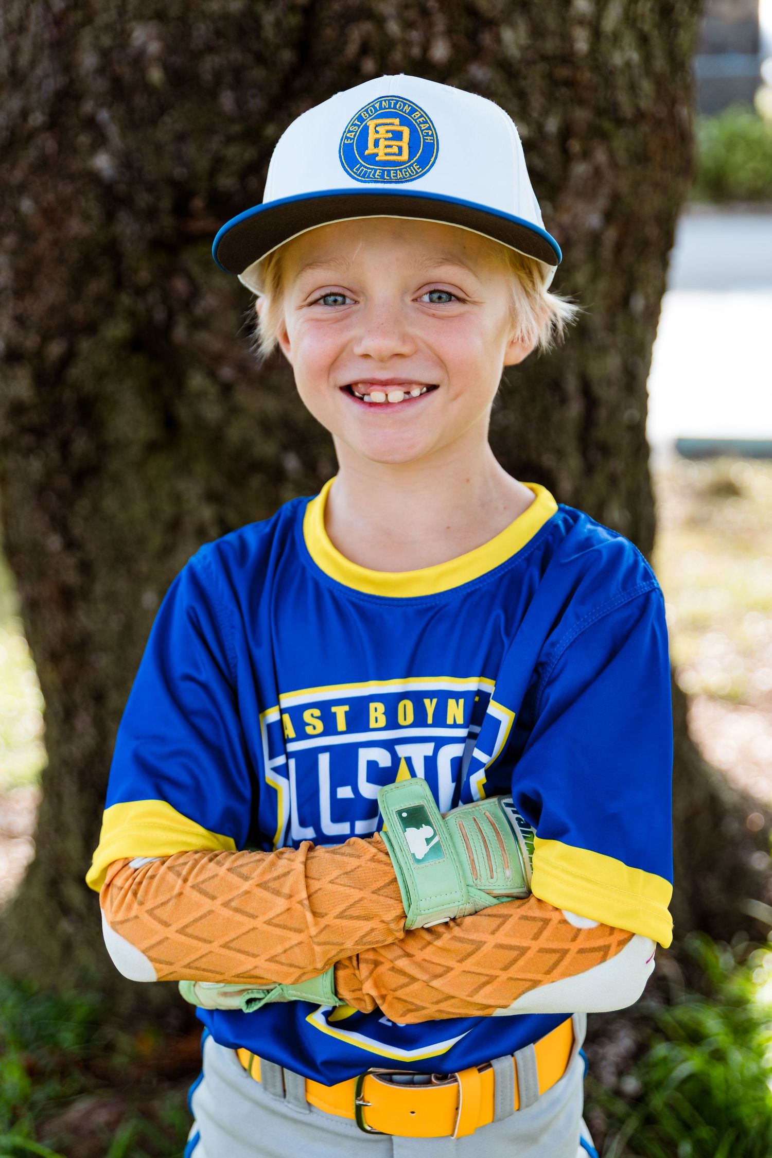 Young baseball player wearing a blue uniform and baseball cap poses outdoors with crossed arms in baseball gloves.