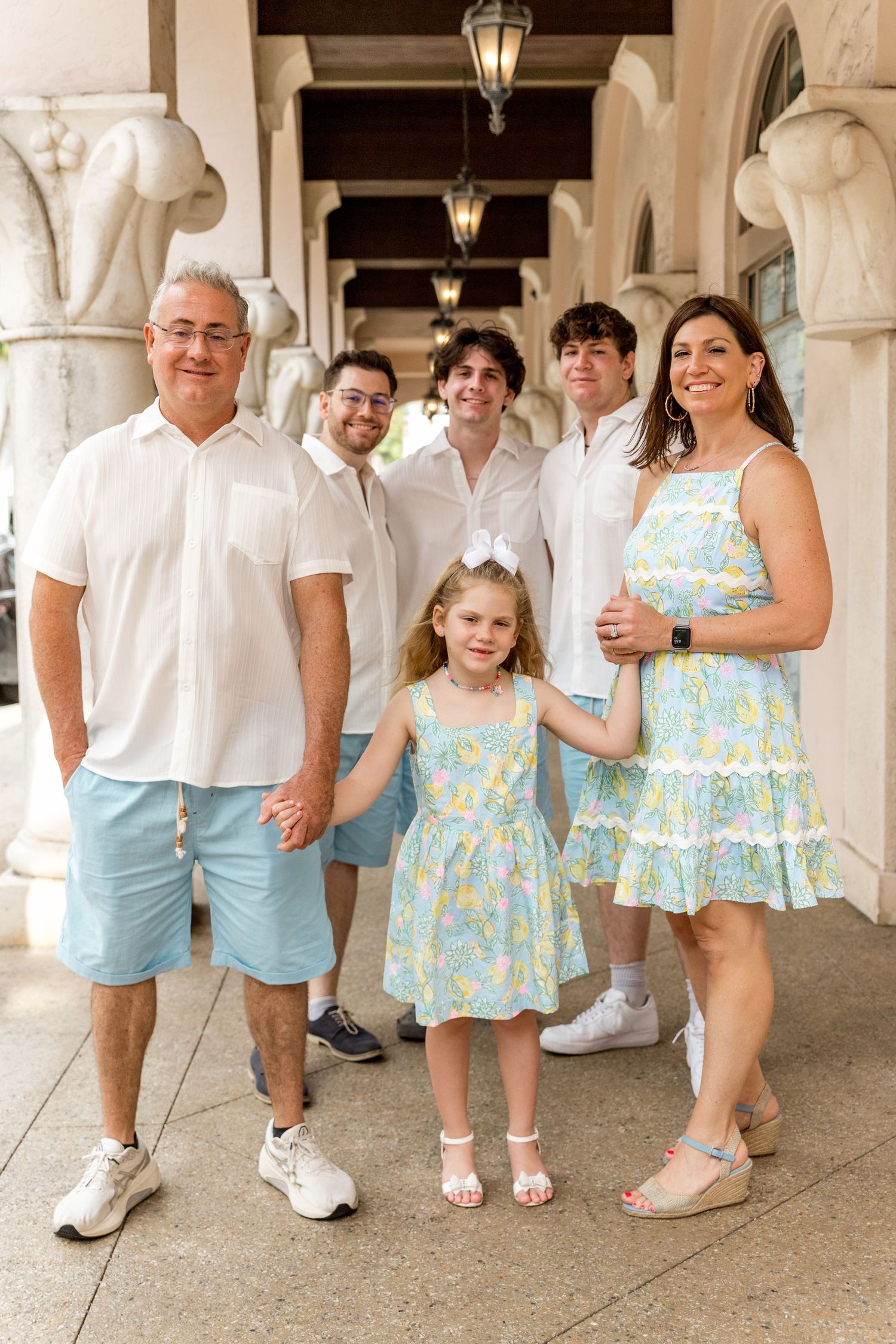 A family group poses in white and light blue outfits under an ornate colonnade with matching mother-daughter dresses.