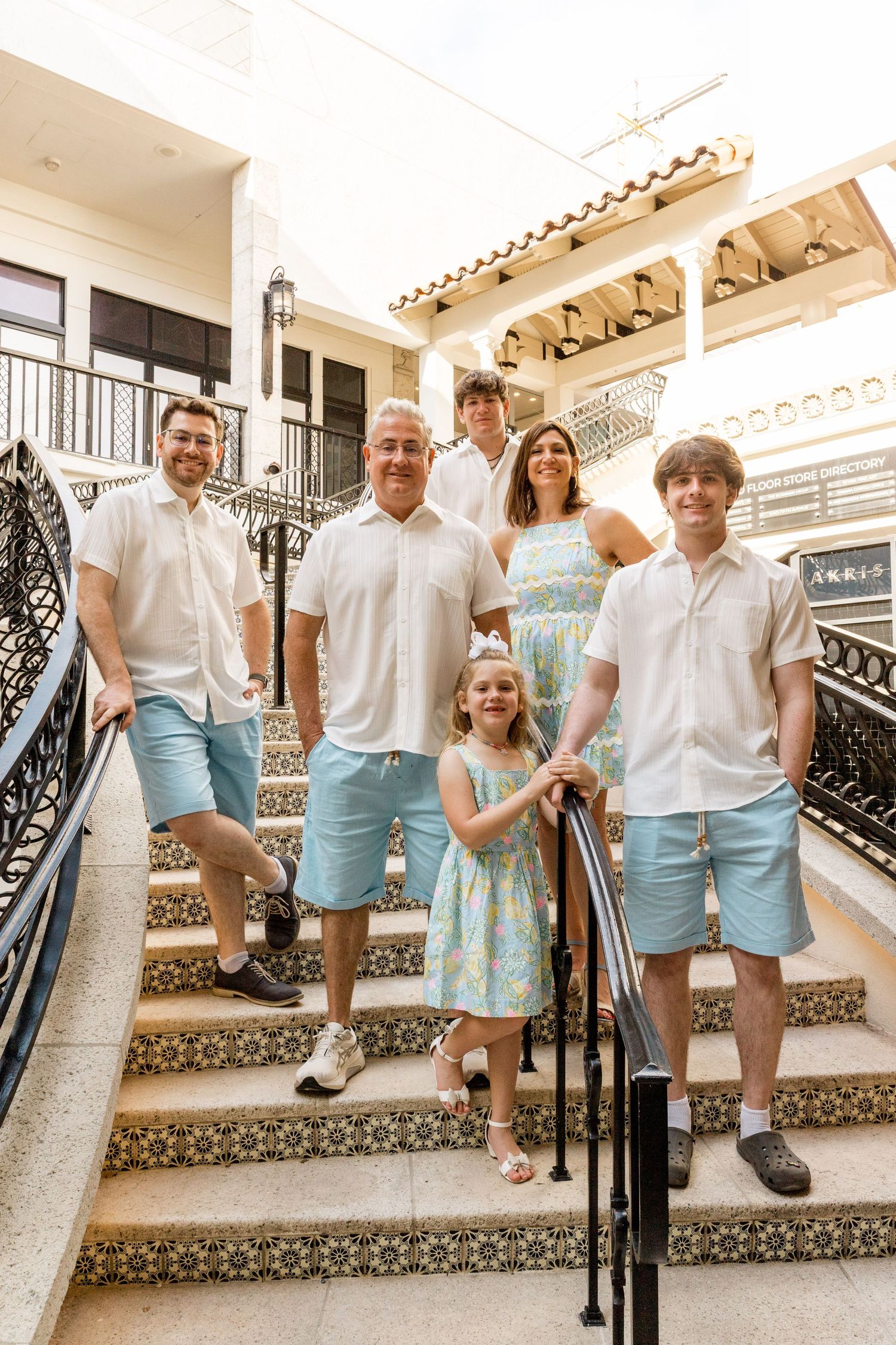 A family in coordinated light blue and white outfits poses on ornate tiled stairs at a Mediterranean-style venue.