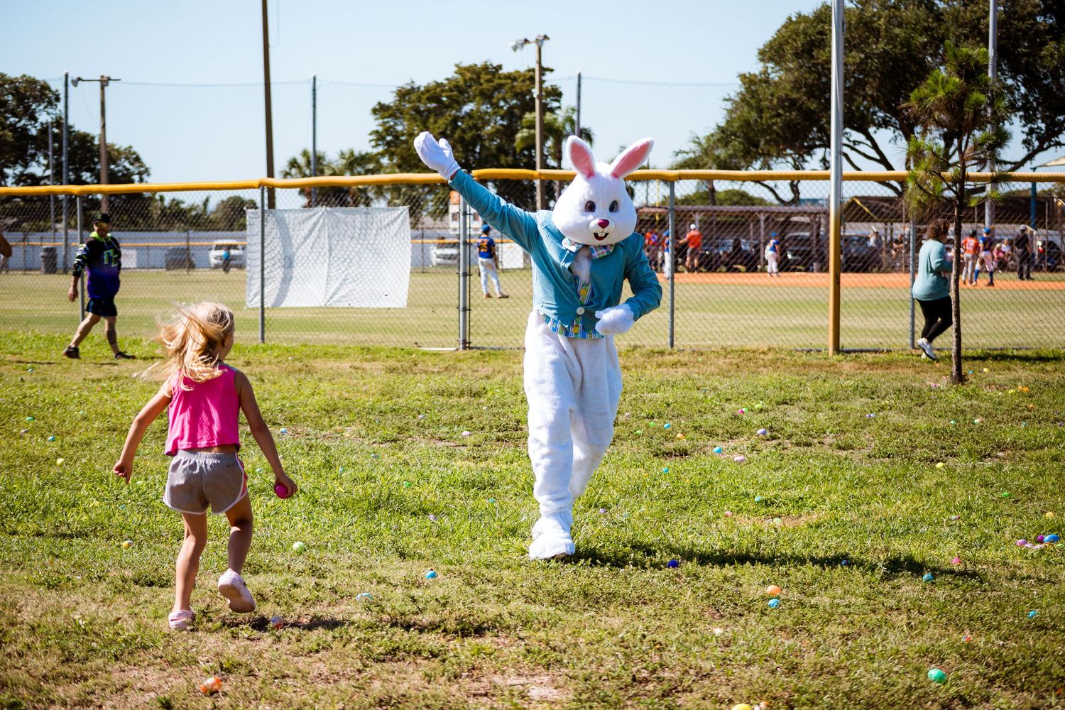 A person in an Easter Bunny costume plays on a sunny field with a small child in pink running nearby.