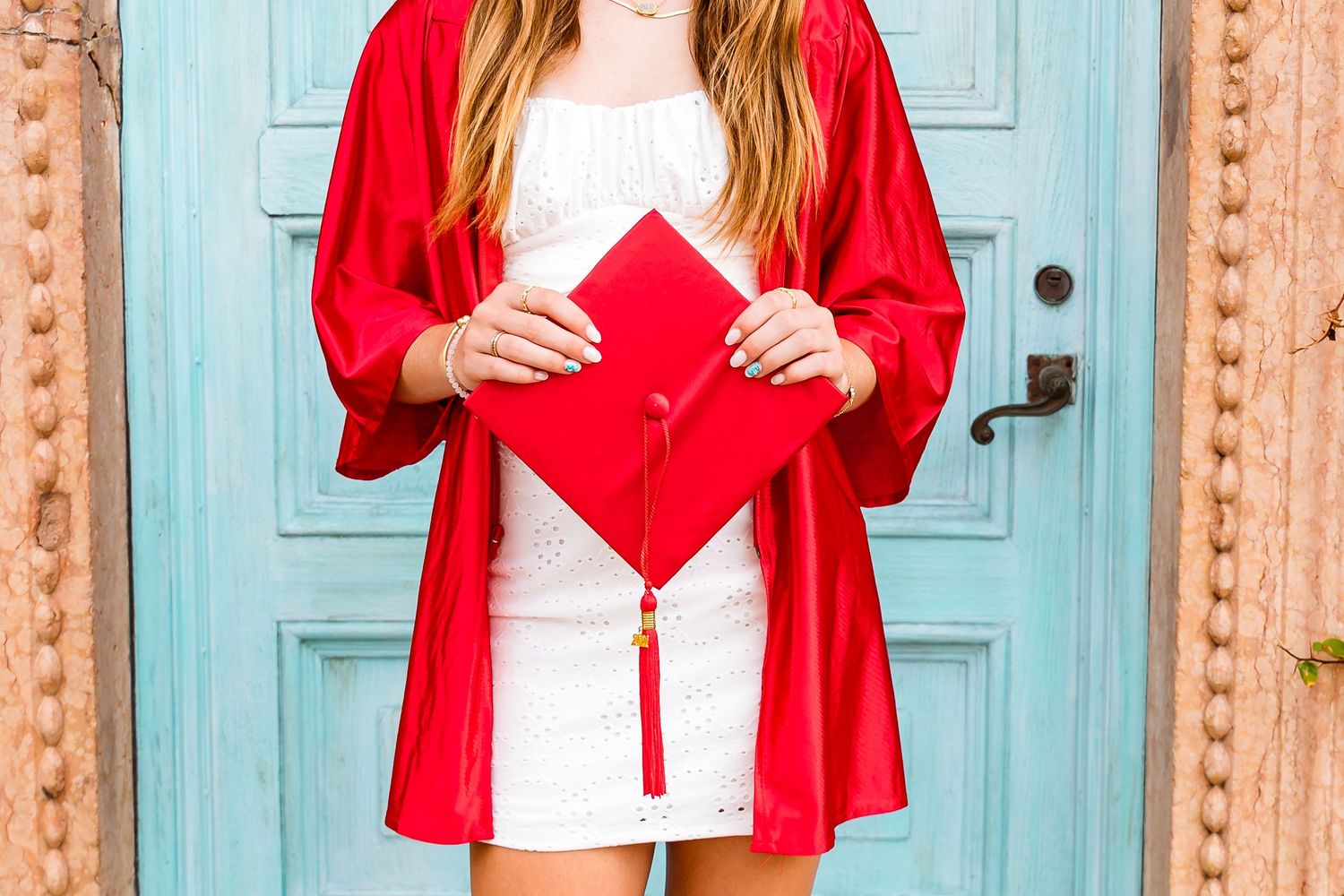 Someone holds a red graduation cap against a white dress and red cardigan in front of a turquoise door.