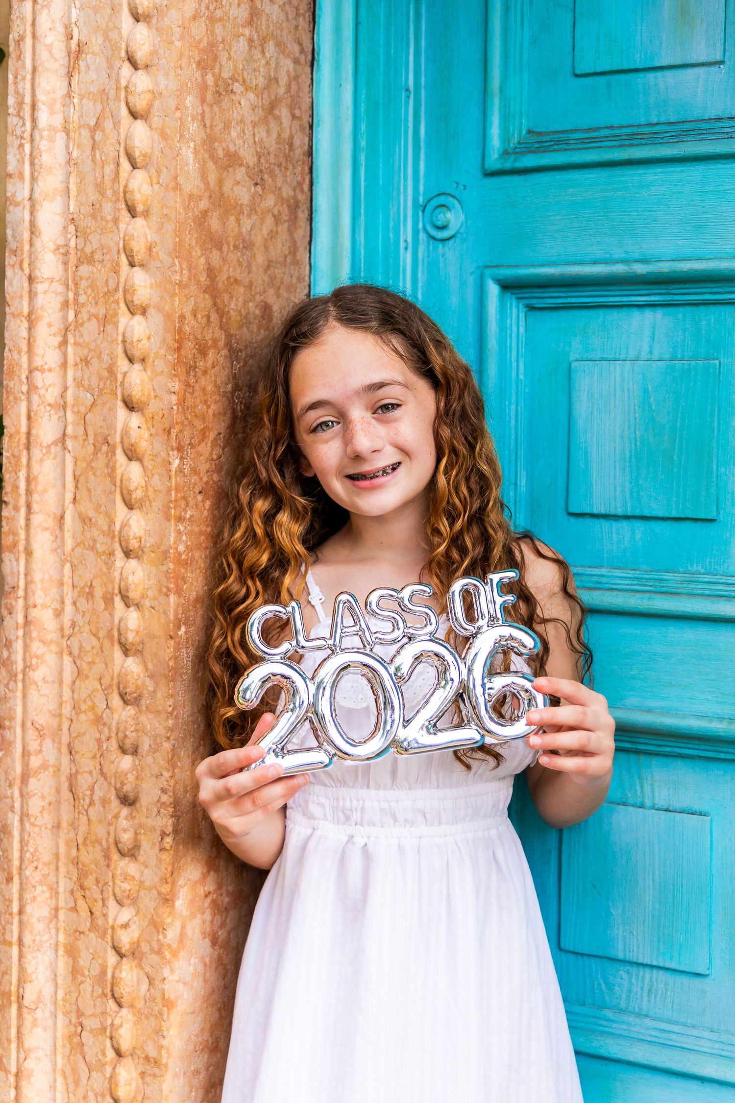 Smiling girl with curly hair in white dress holding Class of 2026 silver balloon sign by turquoise door.