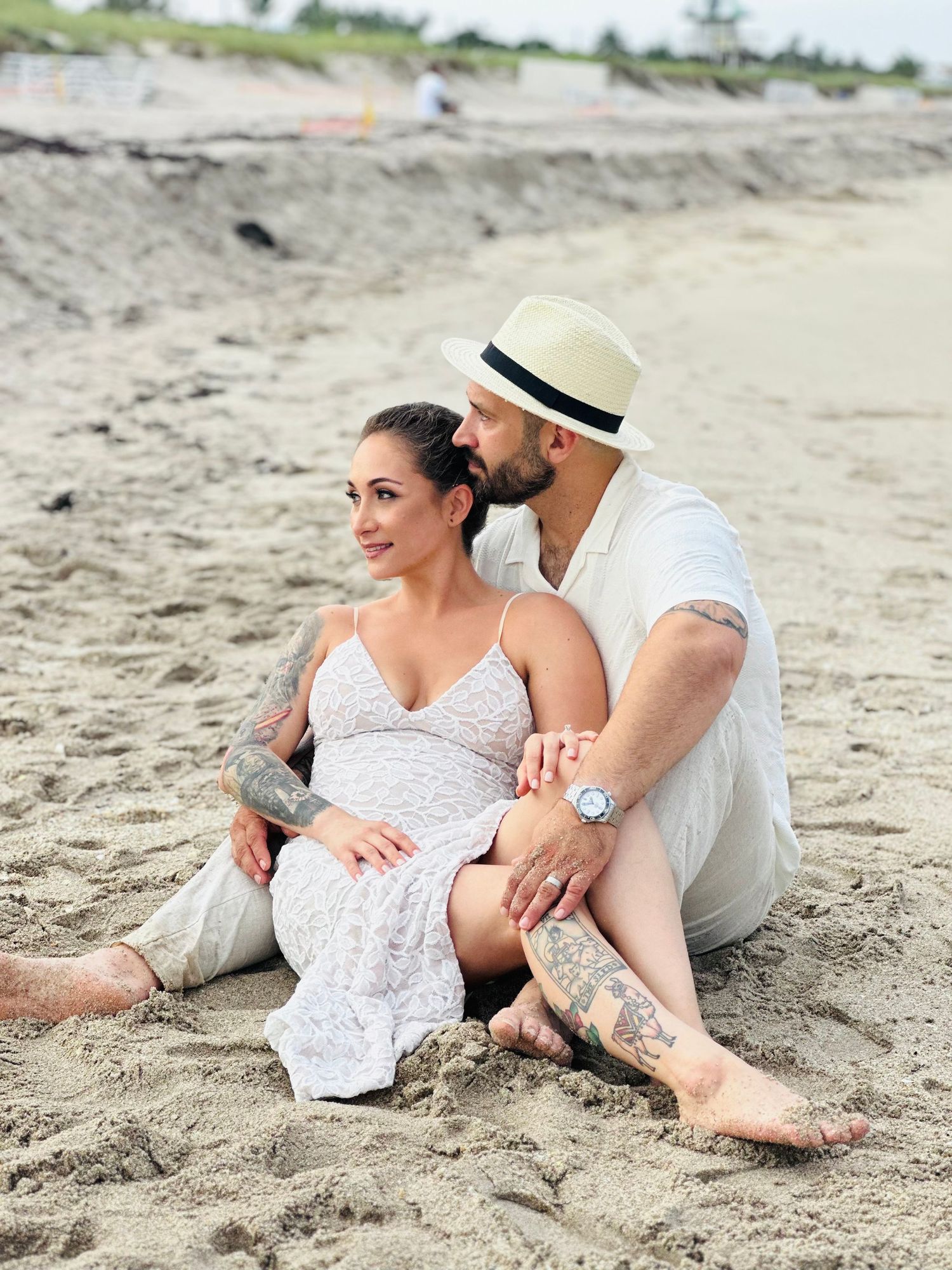 A couple sits intimately on a sandy beach, with one wearing a white dress and the other in a fedora hat and white shirt.