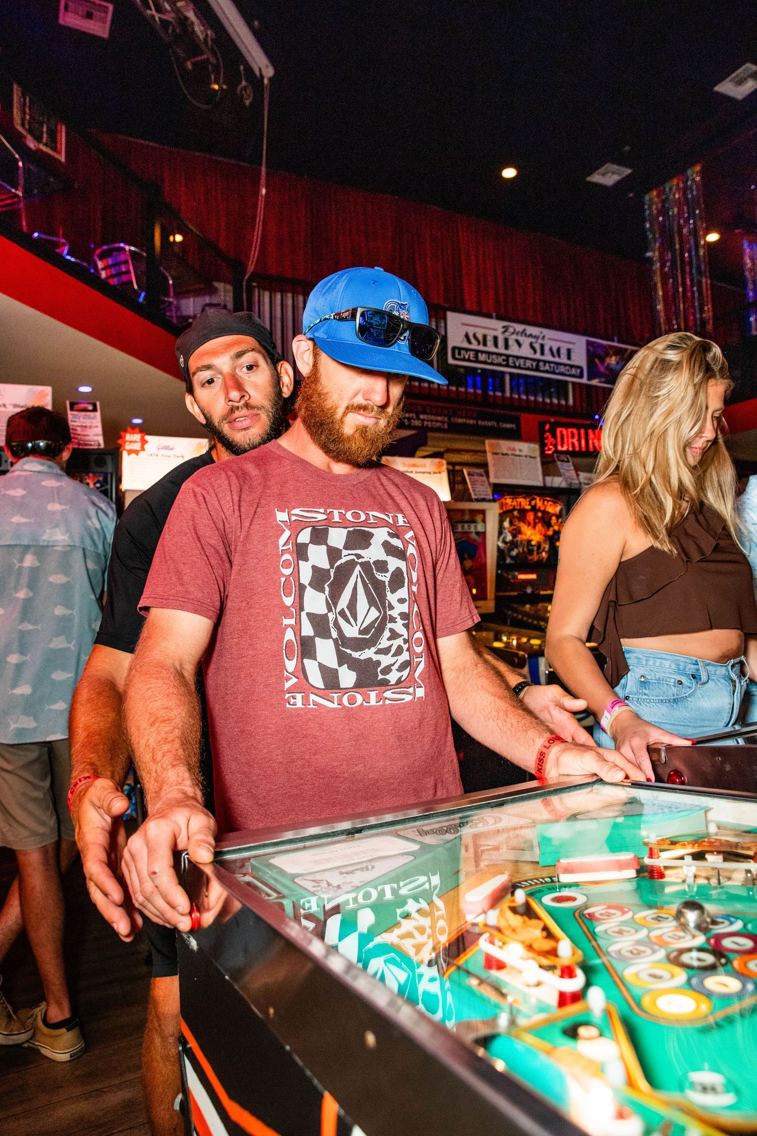 The image shows a group of people playing a pinball machine in a dimly lit, neon-lit arcade or bar setting.