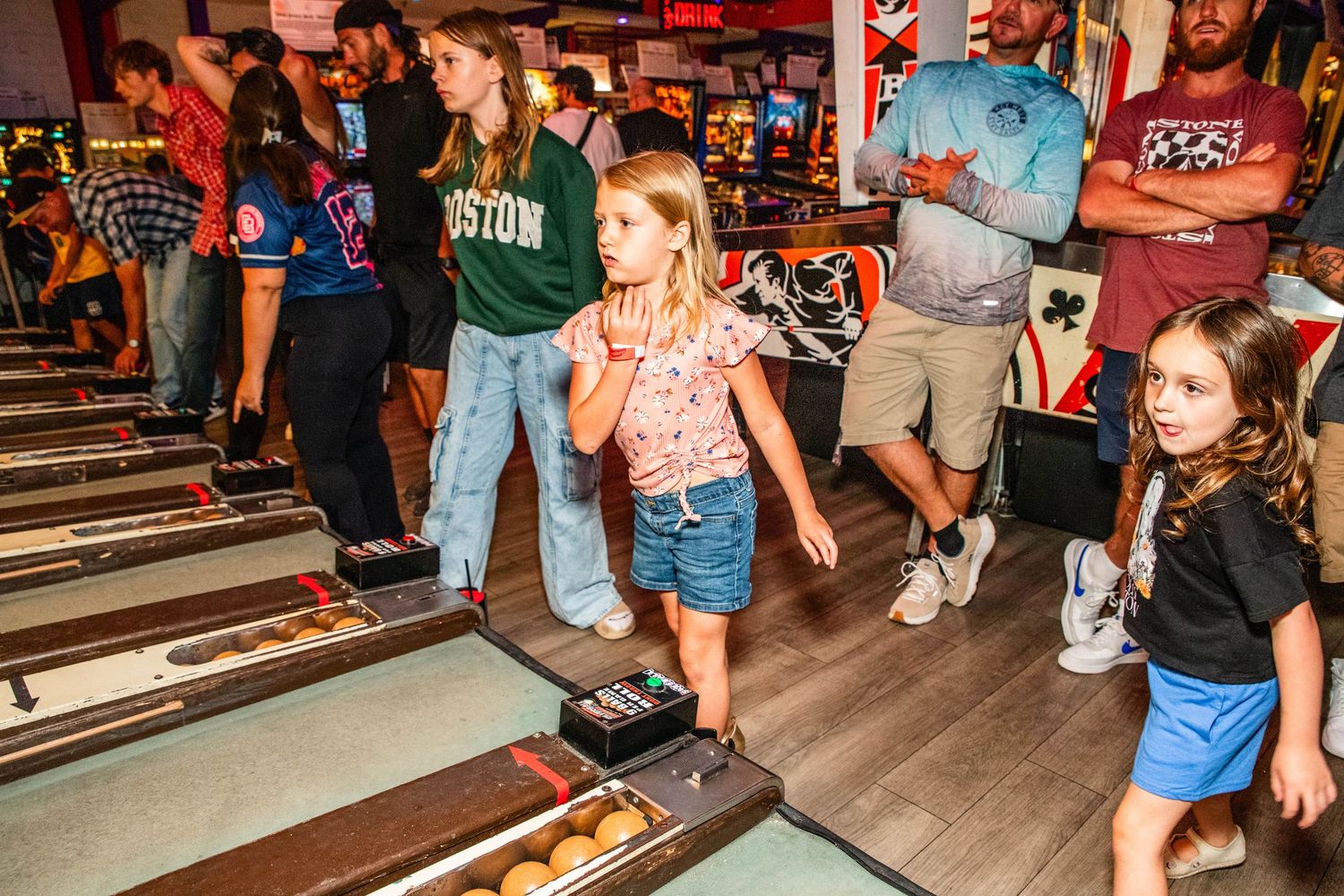 Children playing skee-ball at a lively indoor arcade with adults in background.
