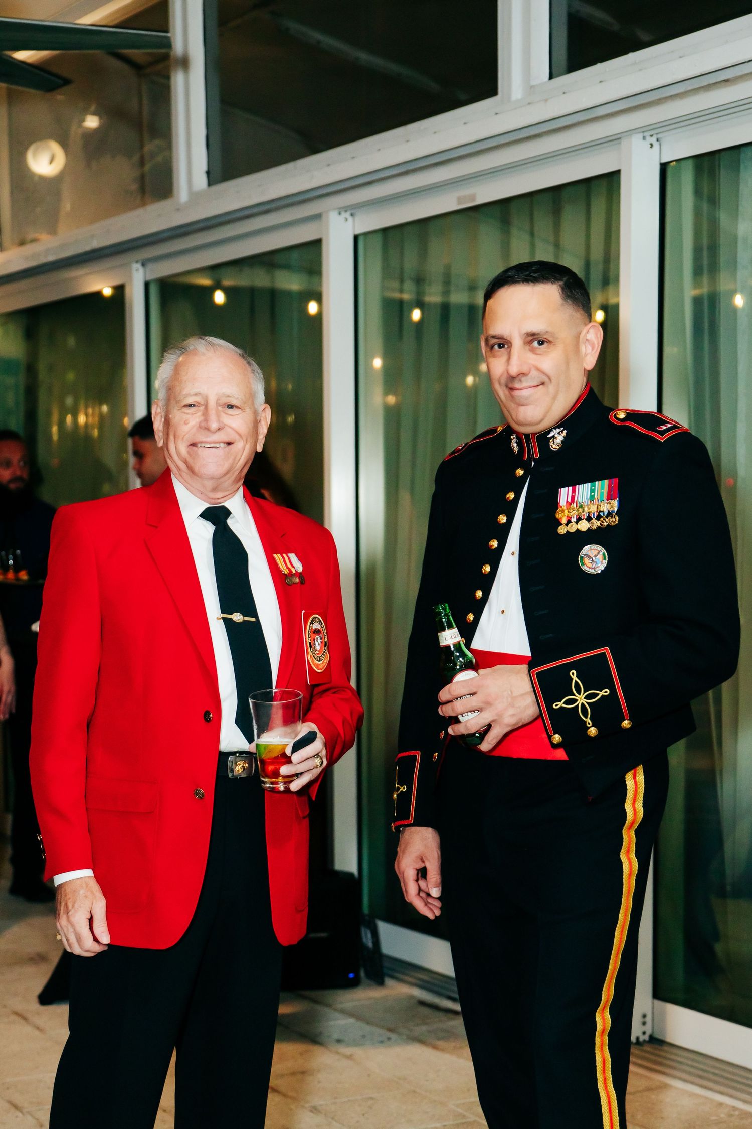 Military veterans in dress uniforms including red jackets and black pants stand together at a formal event.