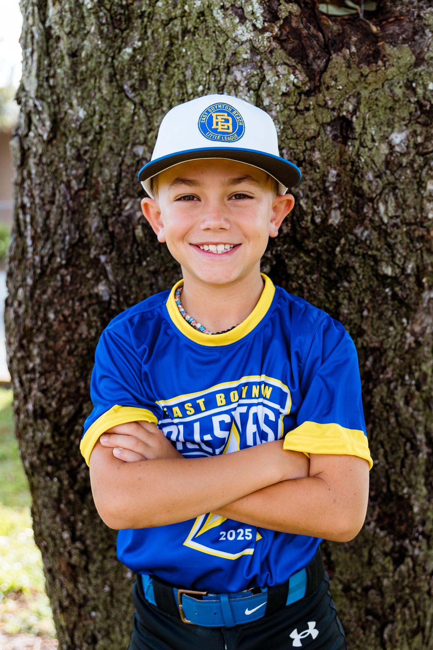 Young baseball player in blue and yellow uniform and baseball cap poses smiling by tree.
