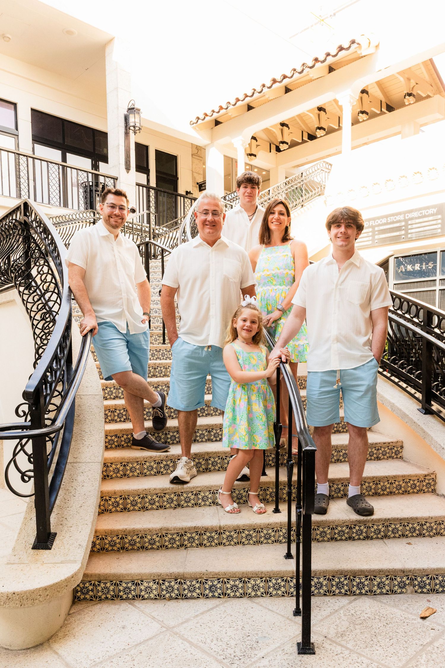 A group in coordinating white and blue outfits poses together on decorative tiled stairs with ornate black railings.