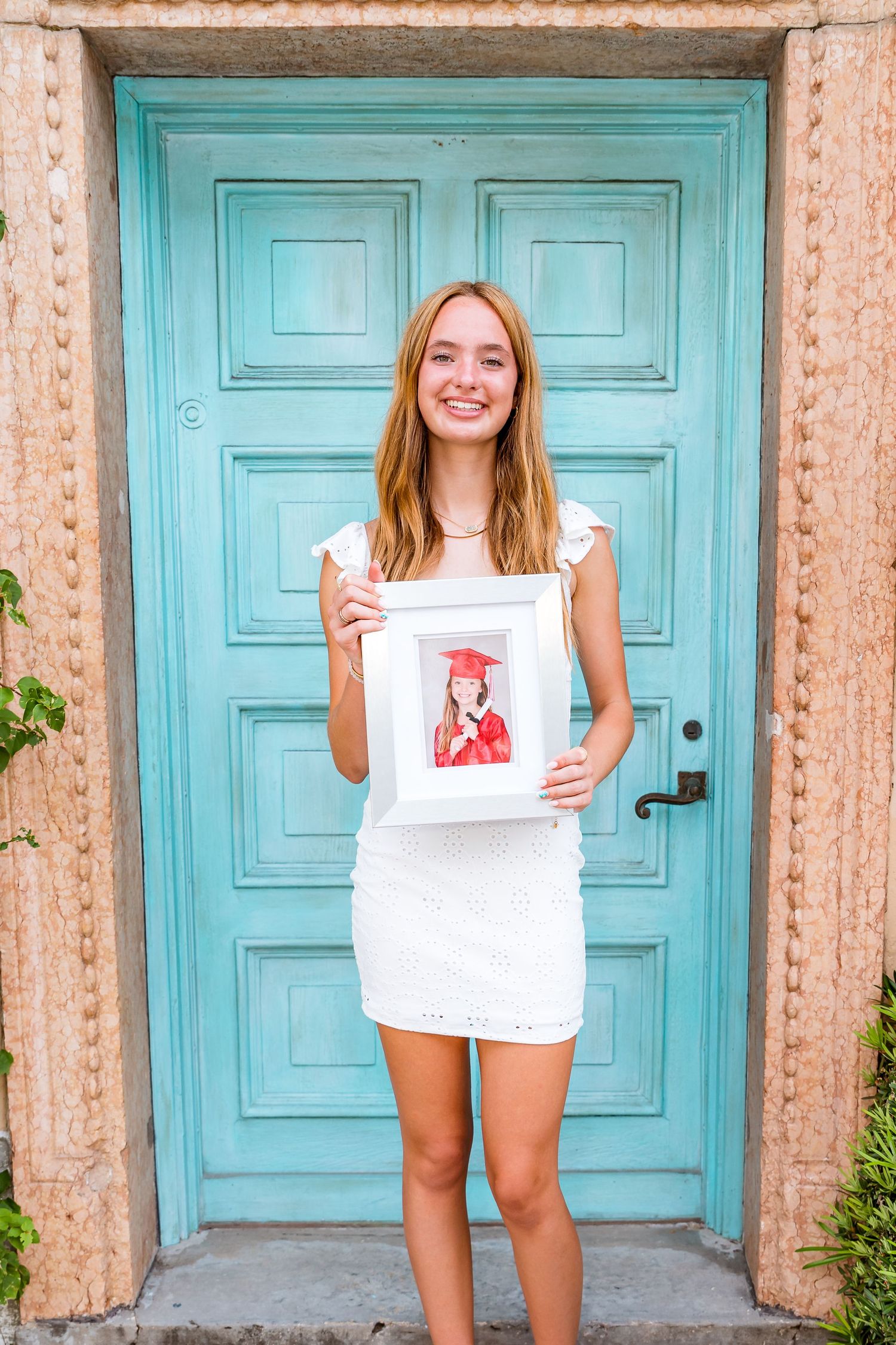 A person in a white dress holds up artwork while standing in front of a turquoise blue door.